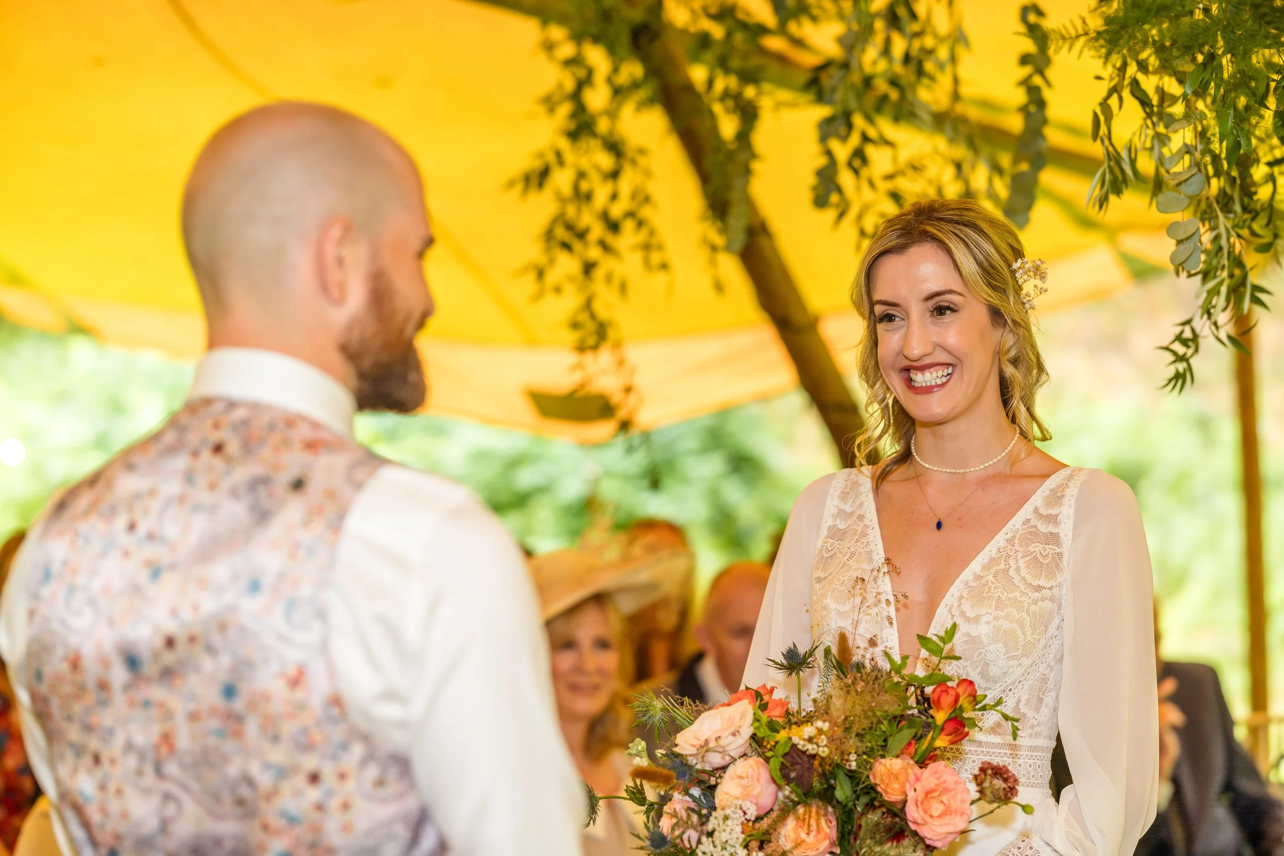 A bride smiling while holding a bouquet of flowers during her wedding ceremony under a yellow canopy