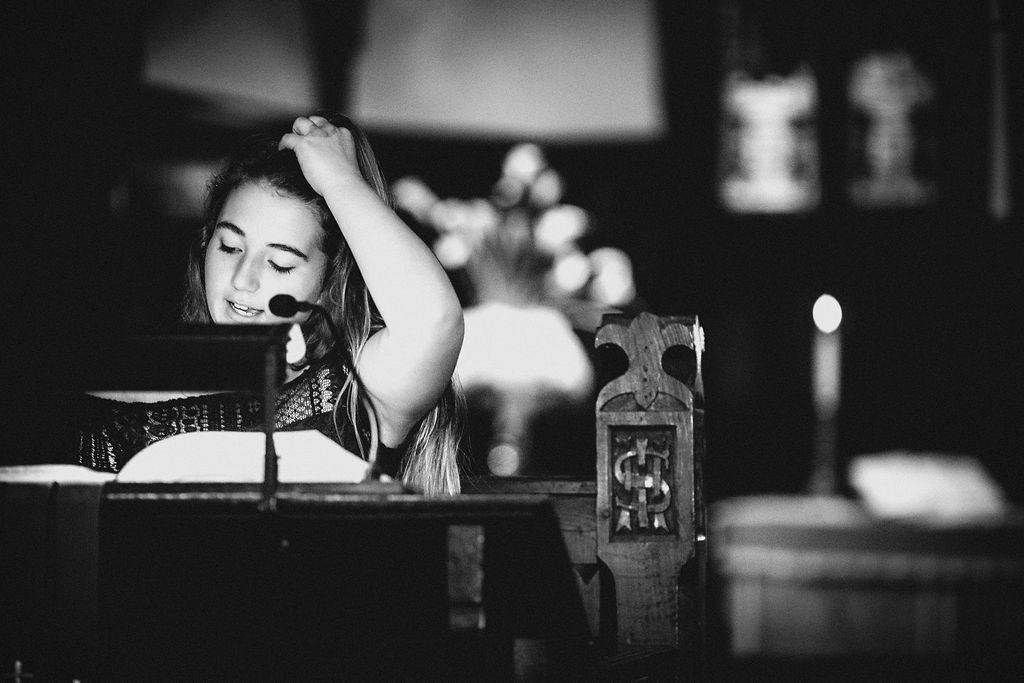 A young girl with long hair is sitting at a piano, holding her head with one hand, and appears to be singing or speaking into a microphone.