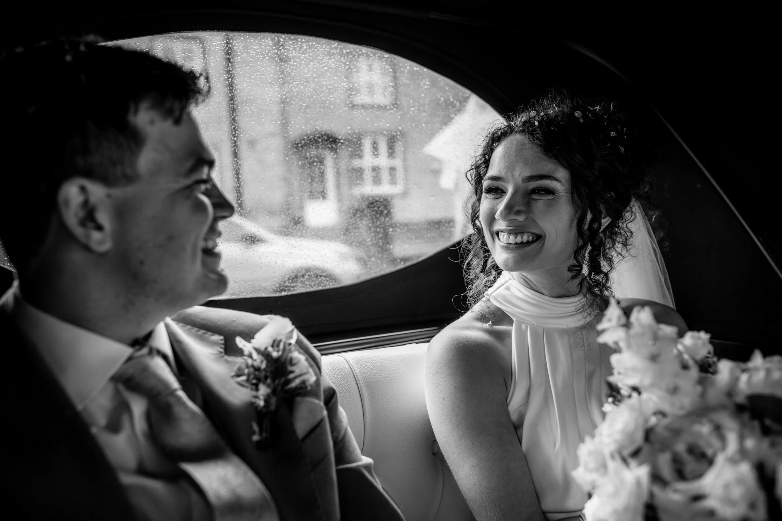 A black and white photo of a bride and groom sitting in the back of a car, smiling at each other during a rainy day.