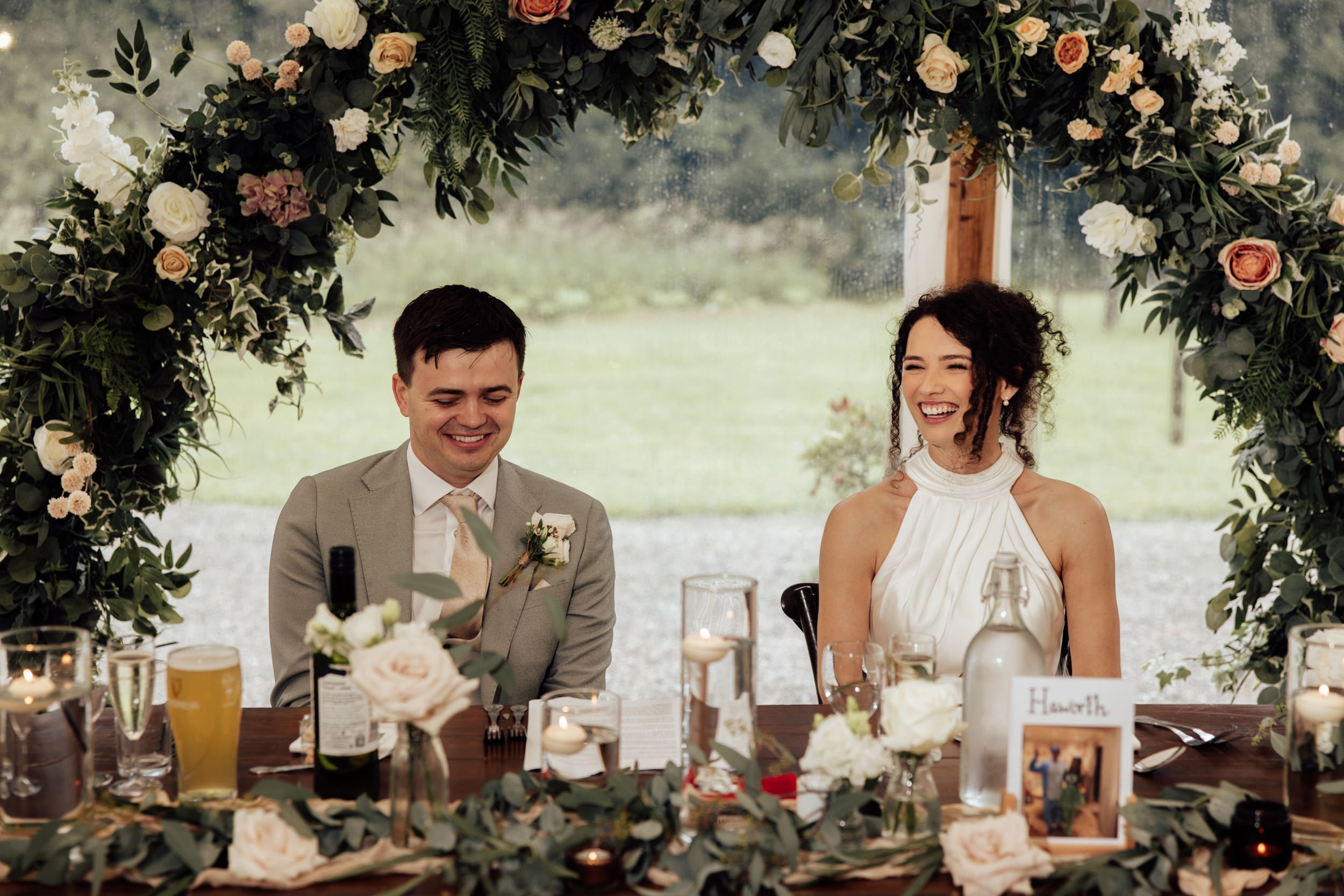 Two people sitting at a decorated wedding table, smiling and laughing, framed by a floral arch with white and blush flowers and greenery.