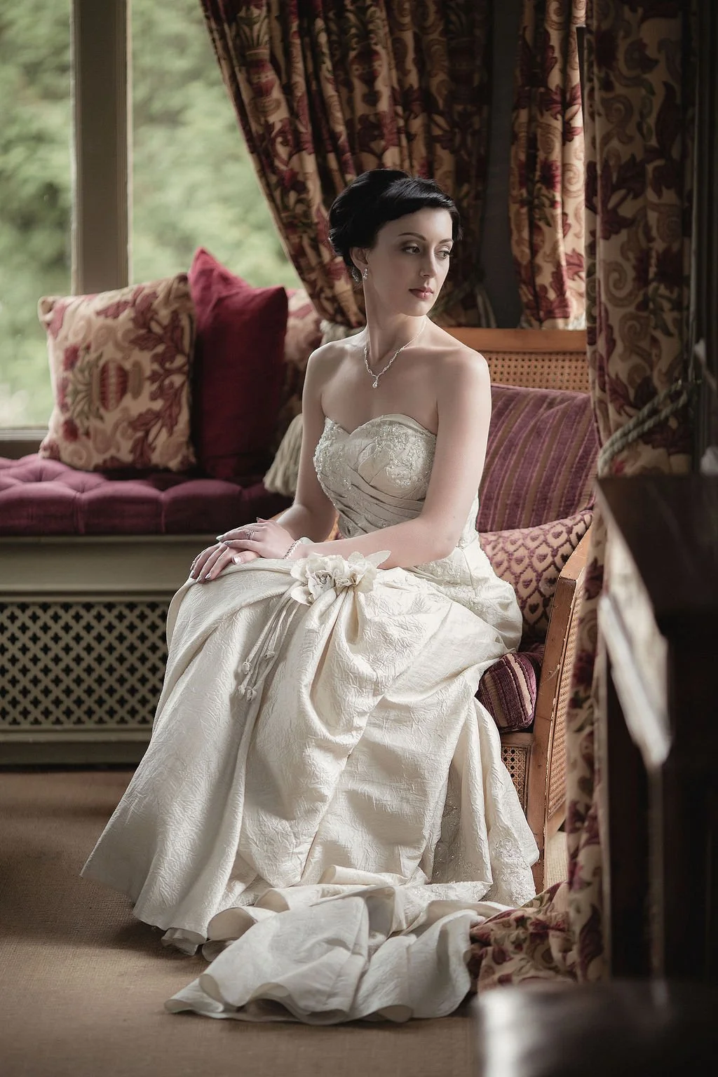 A woman in a wedding dress sitting on a cushioned chair with floral-patterned curtains and pillows in the background.