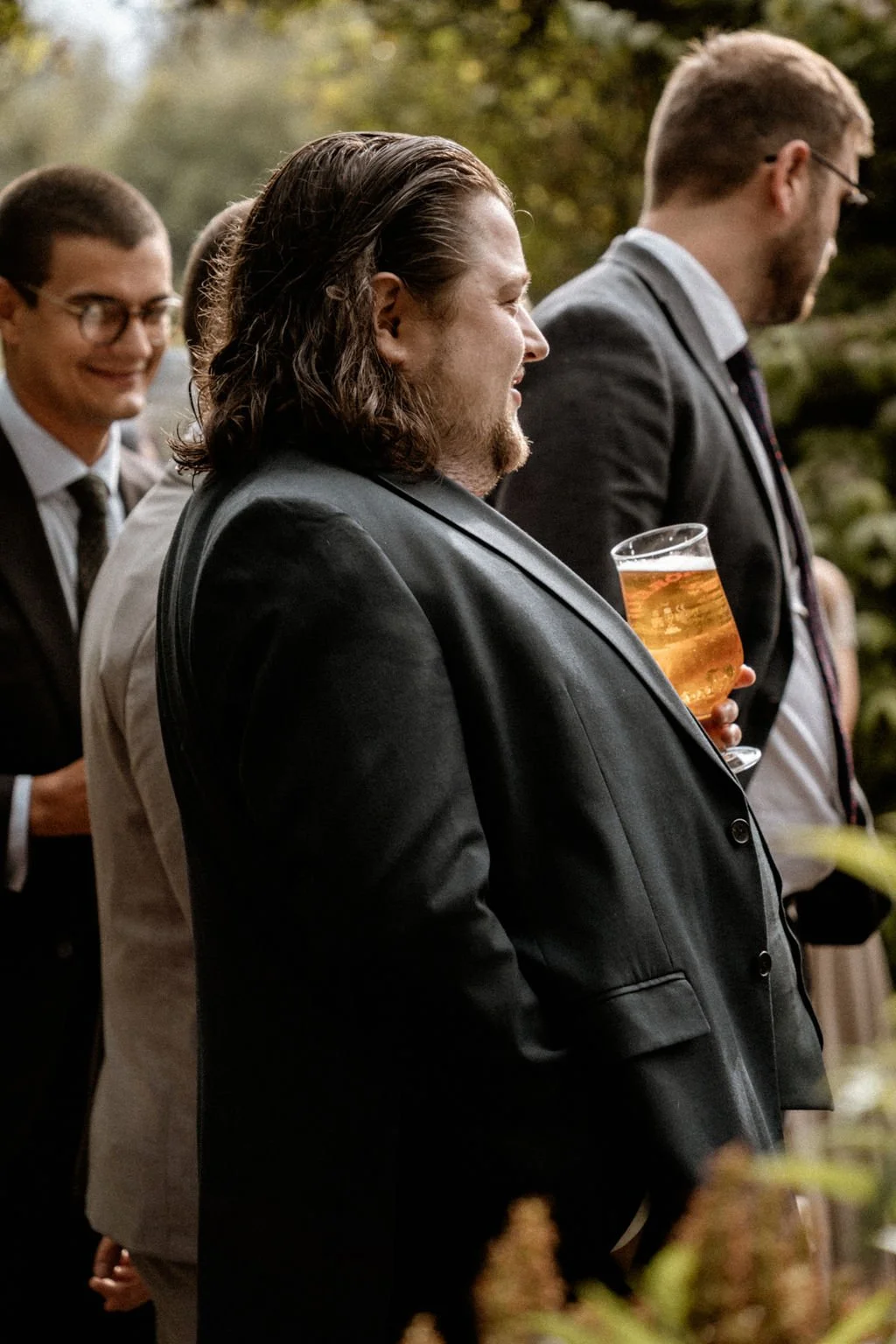 Group of men dressed in suits socializing outdoors, one holding a glass of beer.