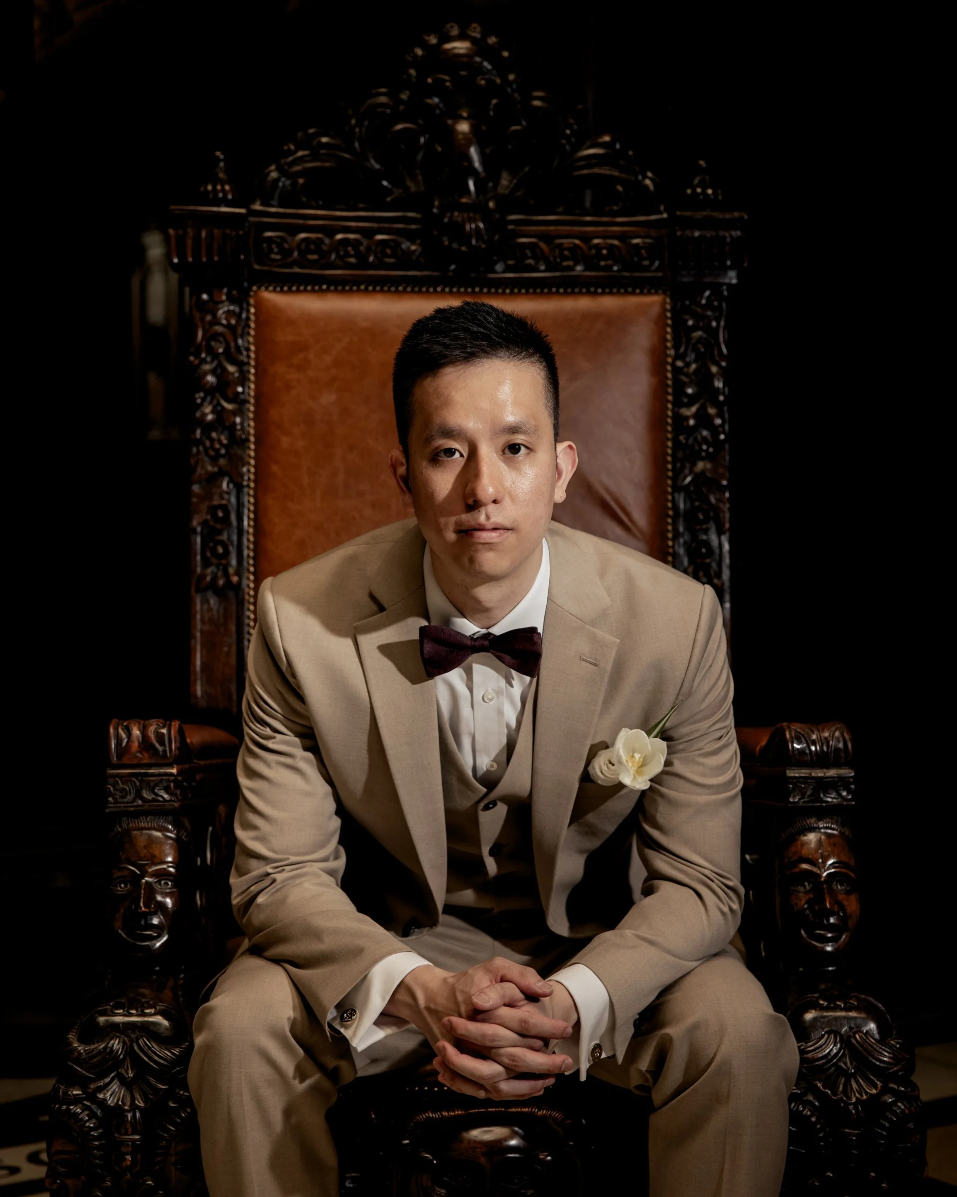 A man in a beige suit and bowtie sitting on a large, ornate wooden throne with intricate carvings, against a dark background.