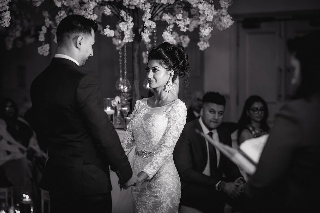 A bride and groom holding hands during their wedding ceremony, surrounded by seated guests, with floral decorations in the background.