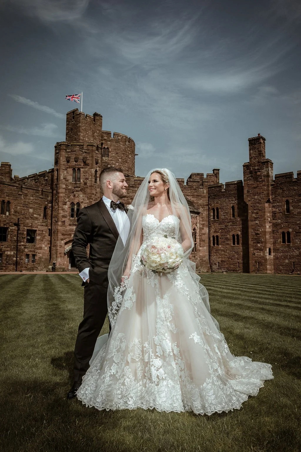 Bride and groom in wedding attire standing in front of a castle, bride holding a bouquet of white roses, bride wearing a lace wedding gown and veil, groom in a black tuxedo and bow tie, castle made of red stone with a flag on top, grassy area in front.