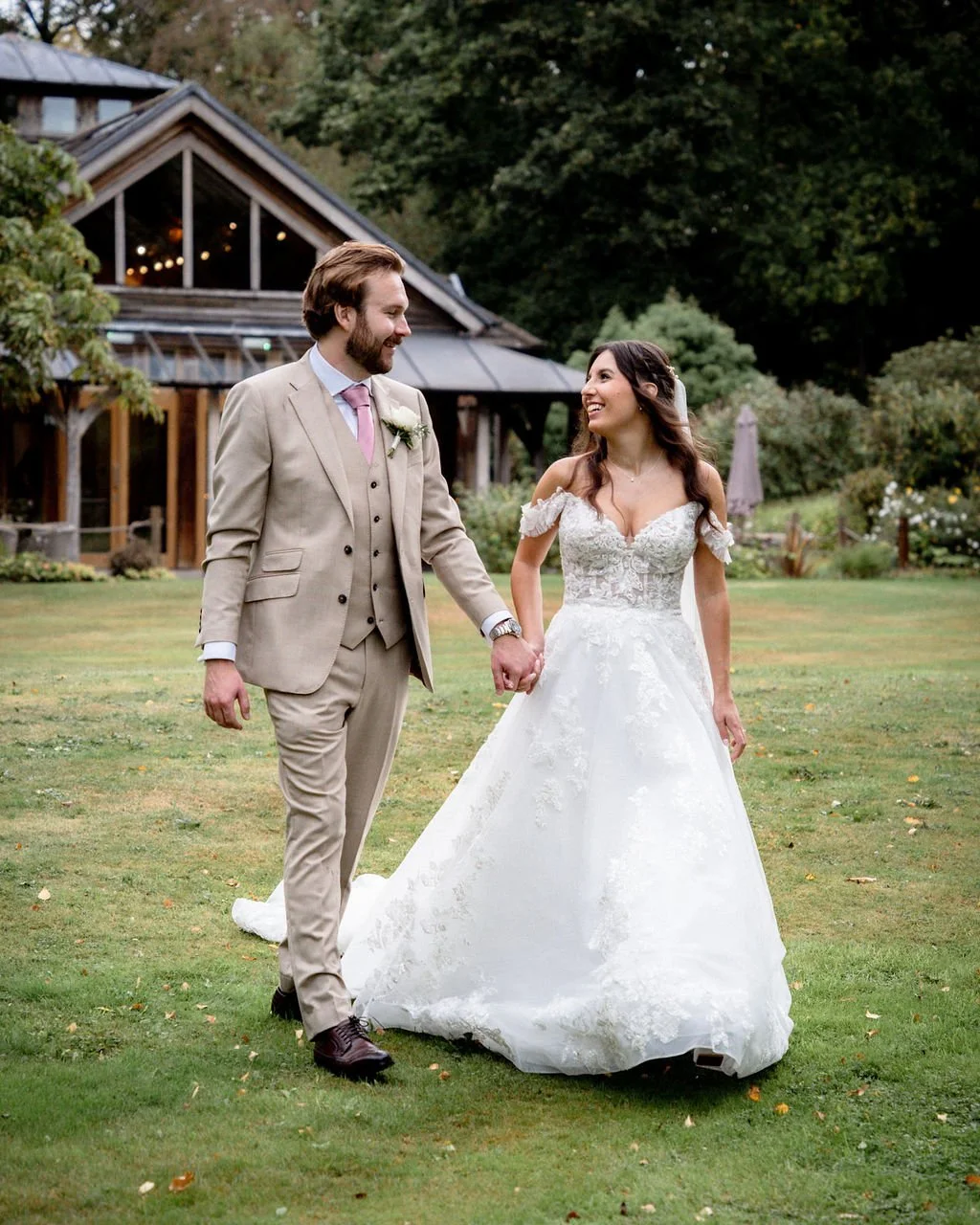 A newlywed couple walking on a grassy area, holding hands and smiling at each other, with a rustic house and trees in the background.