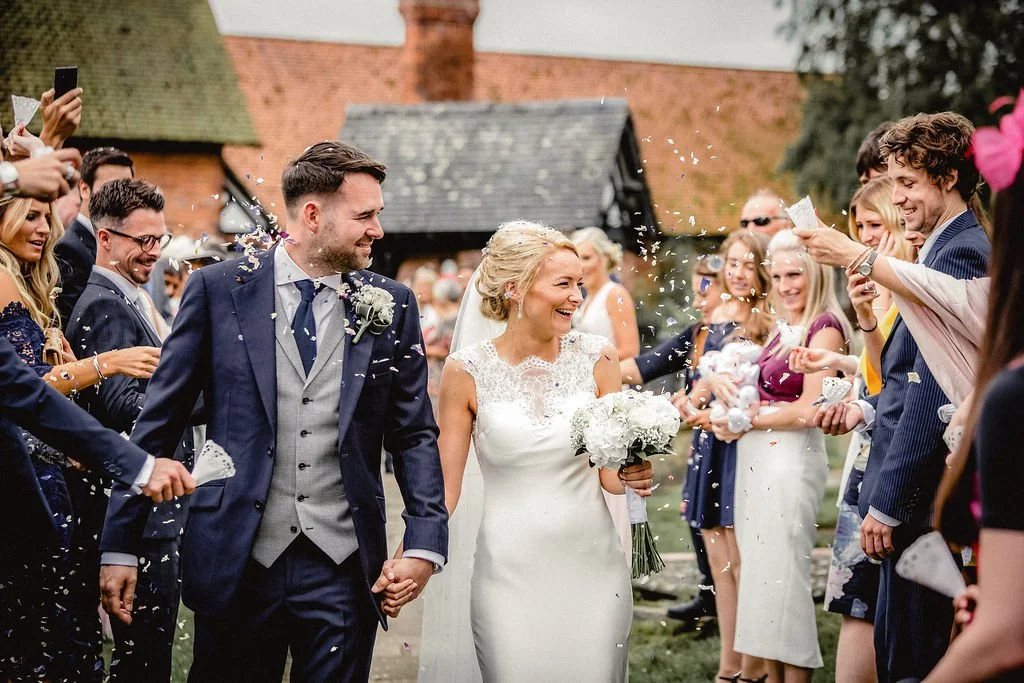 A bride and groom walking hand-in-hand through a crowd of wedding guests outdoors, celebrating with confetti.