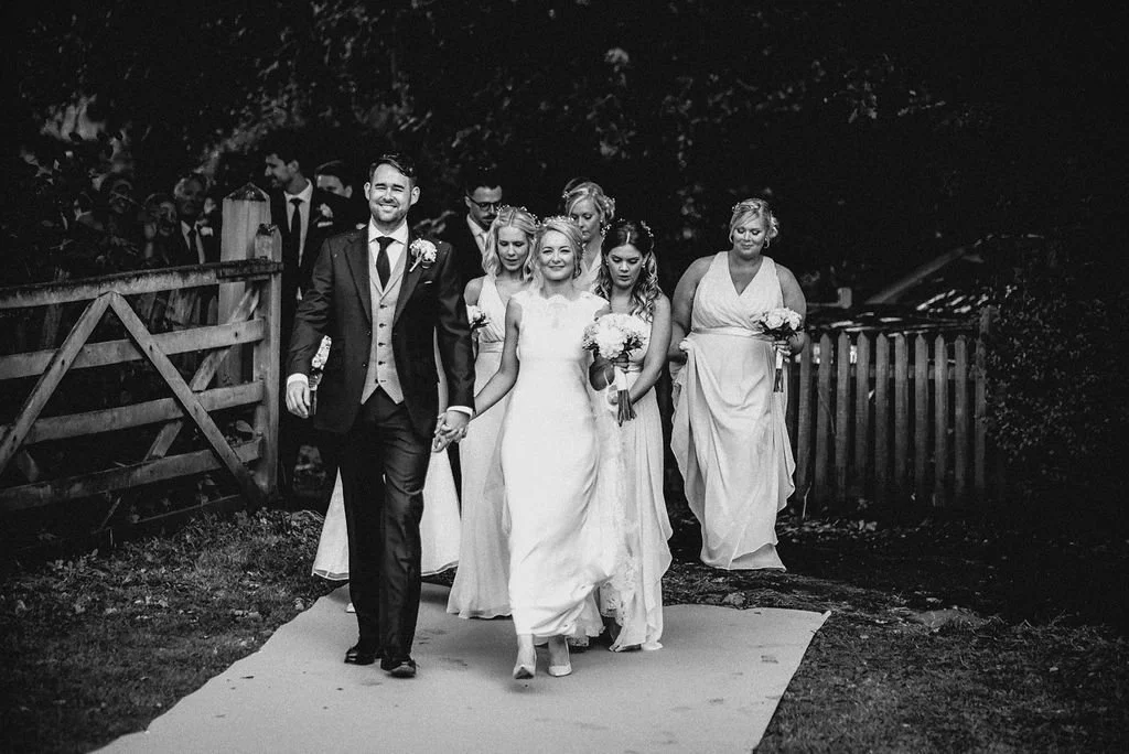 A wedding procession walking along an outdoor path, including a bride in a white dress, a groom in a suit, and bridesmaids. The scene appears to be in the evening or in low light.