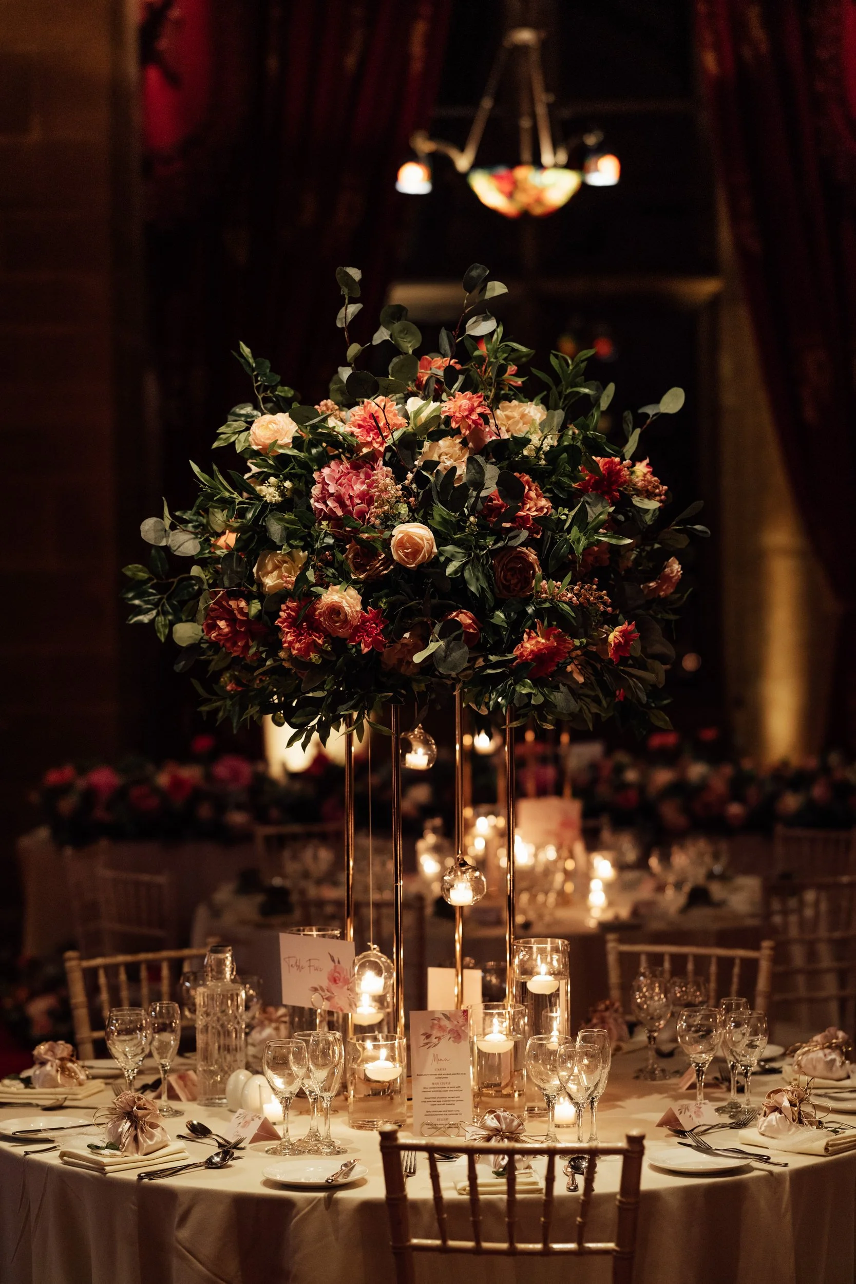 Elegant table setting at a formal event or wedding reception with a tall floral centerpiece, candles, and place settings with glasses and napkins.