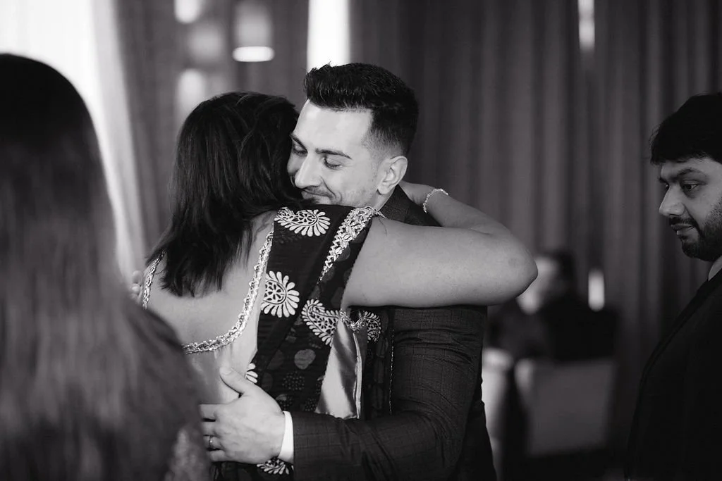 A man and woman hugging at a social gathering, with other people nearby in a room with curtains.