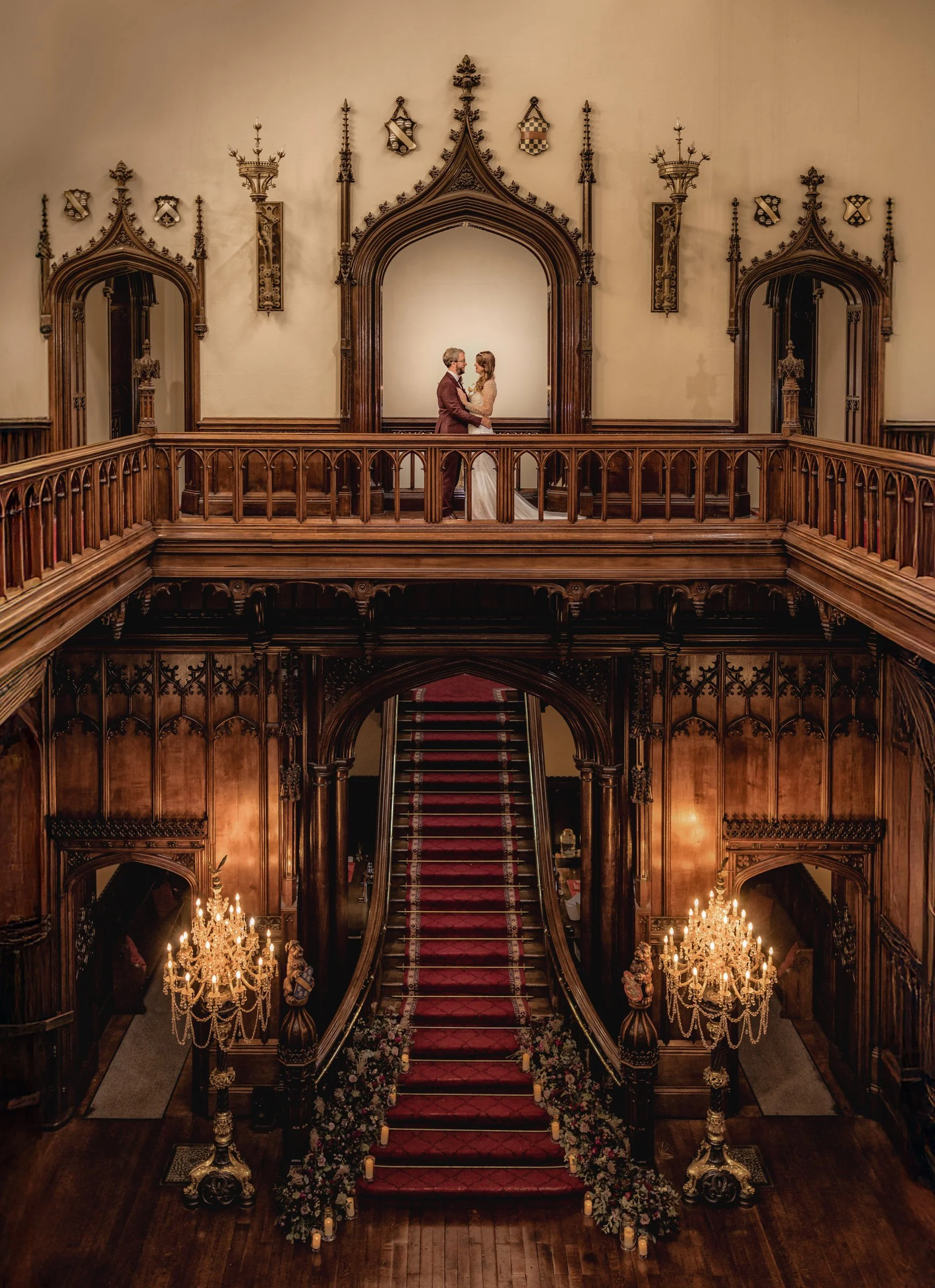 A bride and groom holding hands on a small stage in a grand, historic hall with wood-paneled walls, archways, chandeliers, and a red-carpeted staircase decorated with flowers and candles.