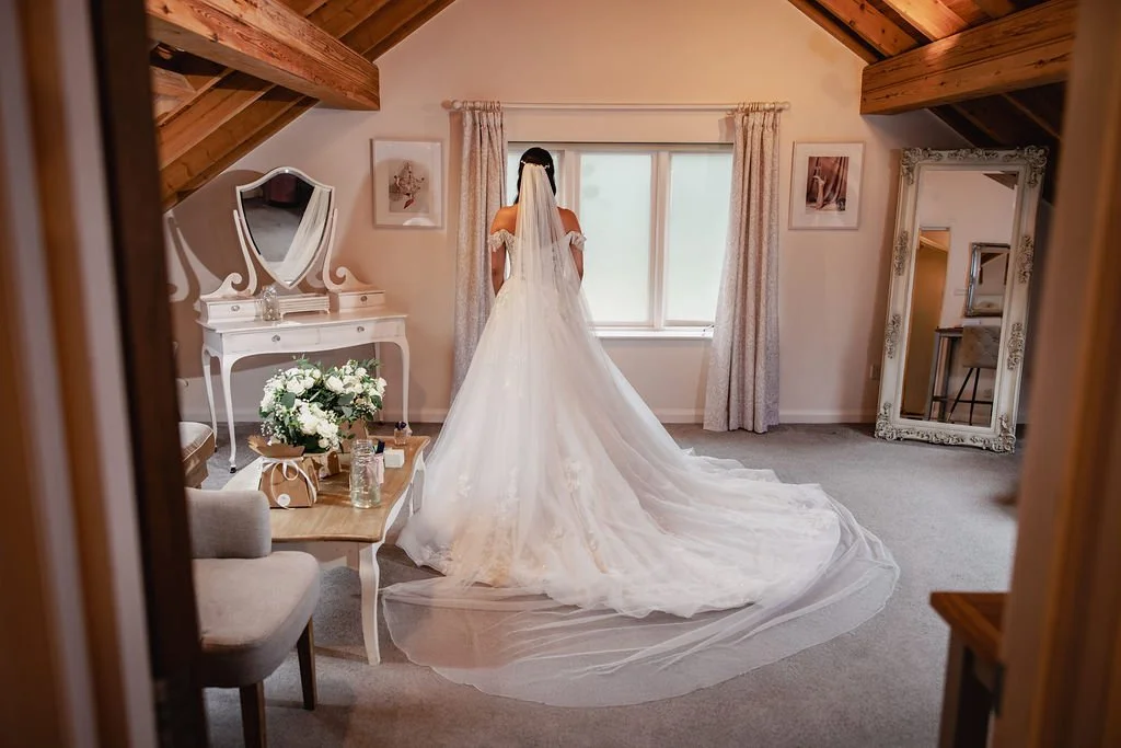 A bride in a wedding gown with a long train, facing a window in a well-lit bedroom with wooden ceiling beams, decorated with framed pictures and a full-length mirror.