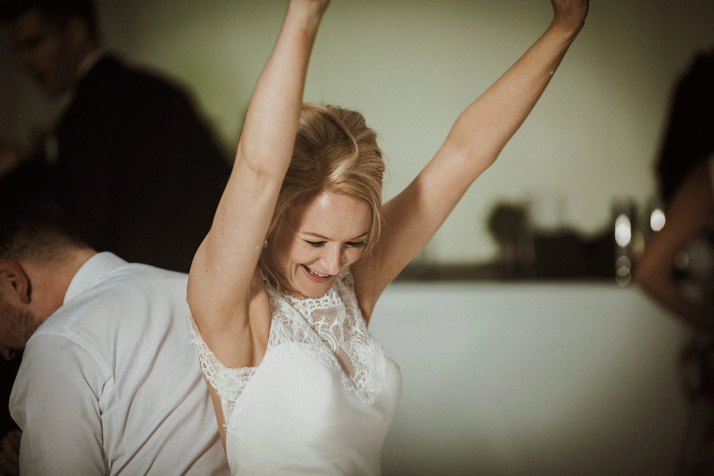 A joyful woman with her arms raised, smiling with closed eyes, wearing a white lace dress, with a man behind her in a light-colored shirt.