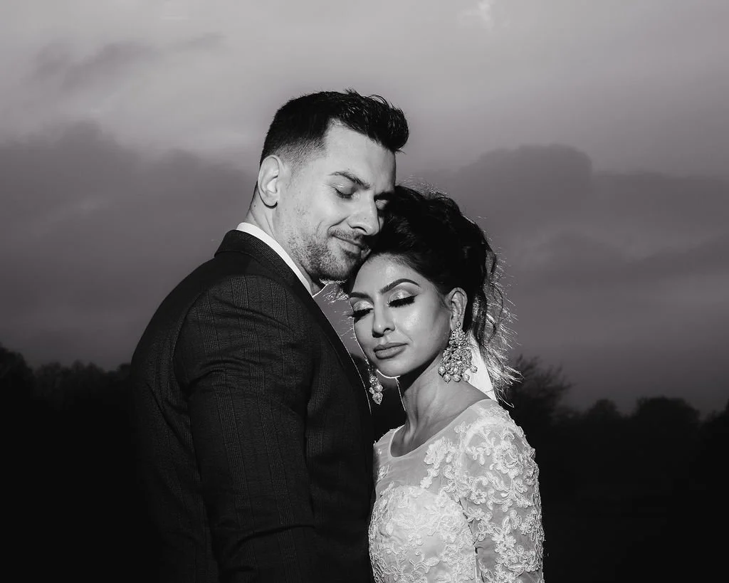 A black and white photo of a couple embracing outdoors, with a cloudy sky in the background. The man is dressed in a suit, and the woman is wearing a lace wedding dress and large earrings, with their eyes closed.