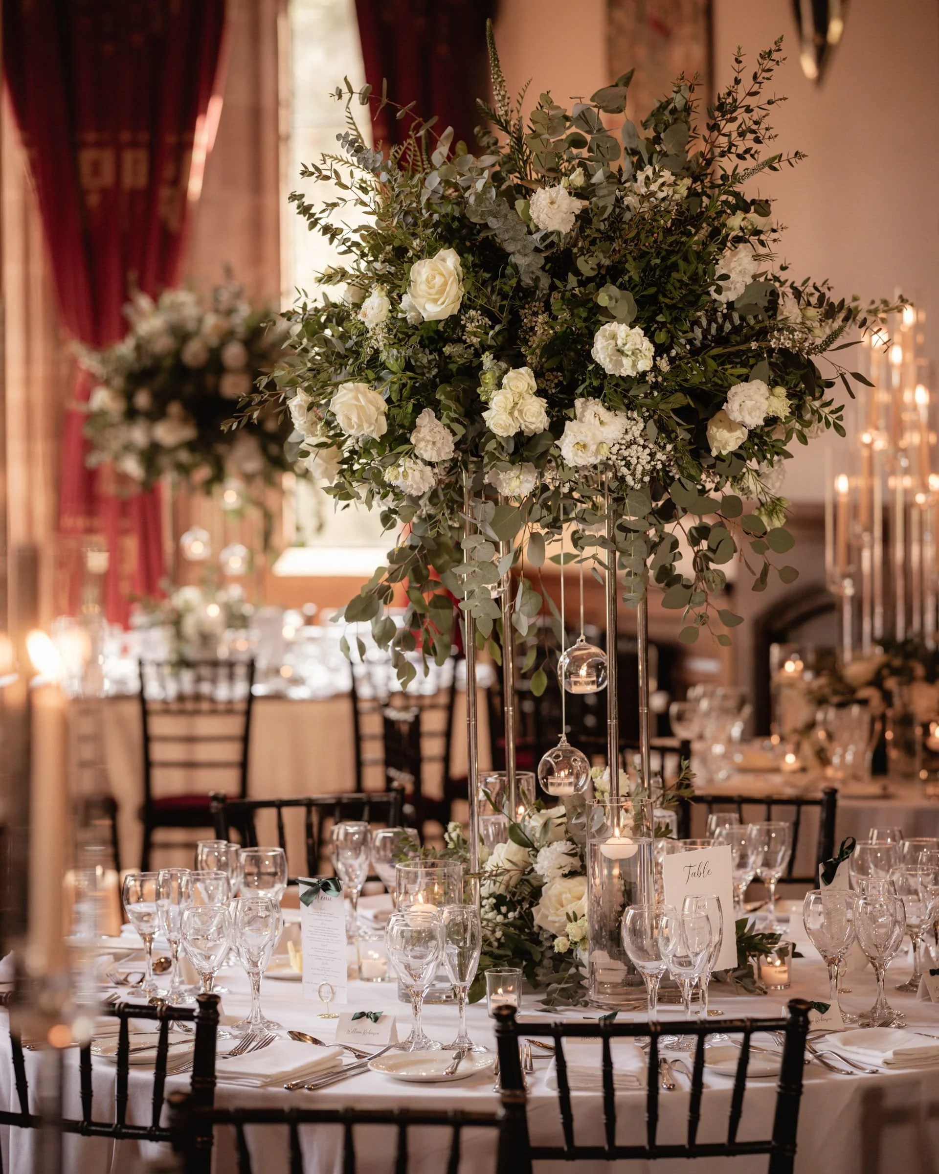 Elegant indoor wedding reception with a tall floral centerpiece of white roses, hydrangeas, and greenery on a table set with glasses and place cards, soft warm lighting, and red curtains in the background.