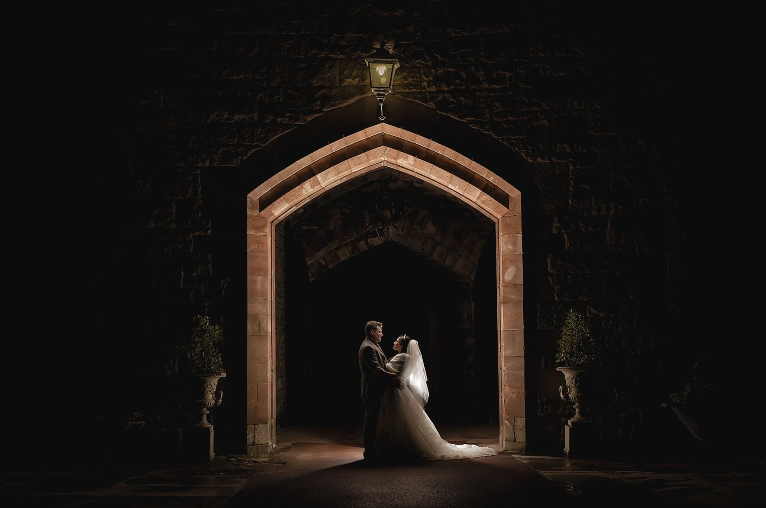 A bride and groom holding hands and gazing at each other at night under an illuminated stone archway, with potted plants on both sides.