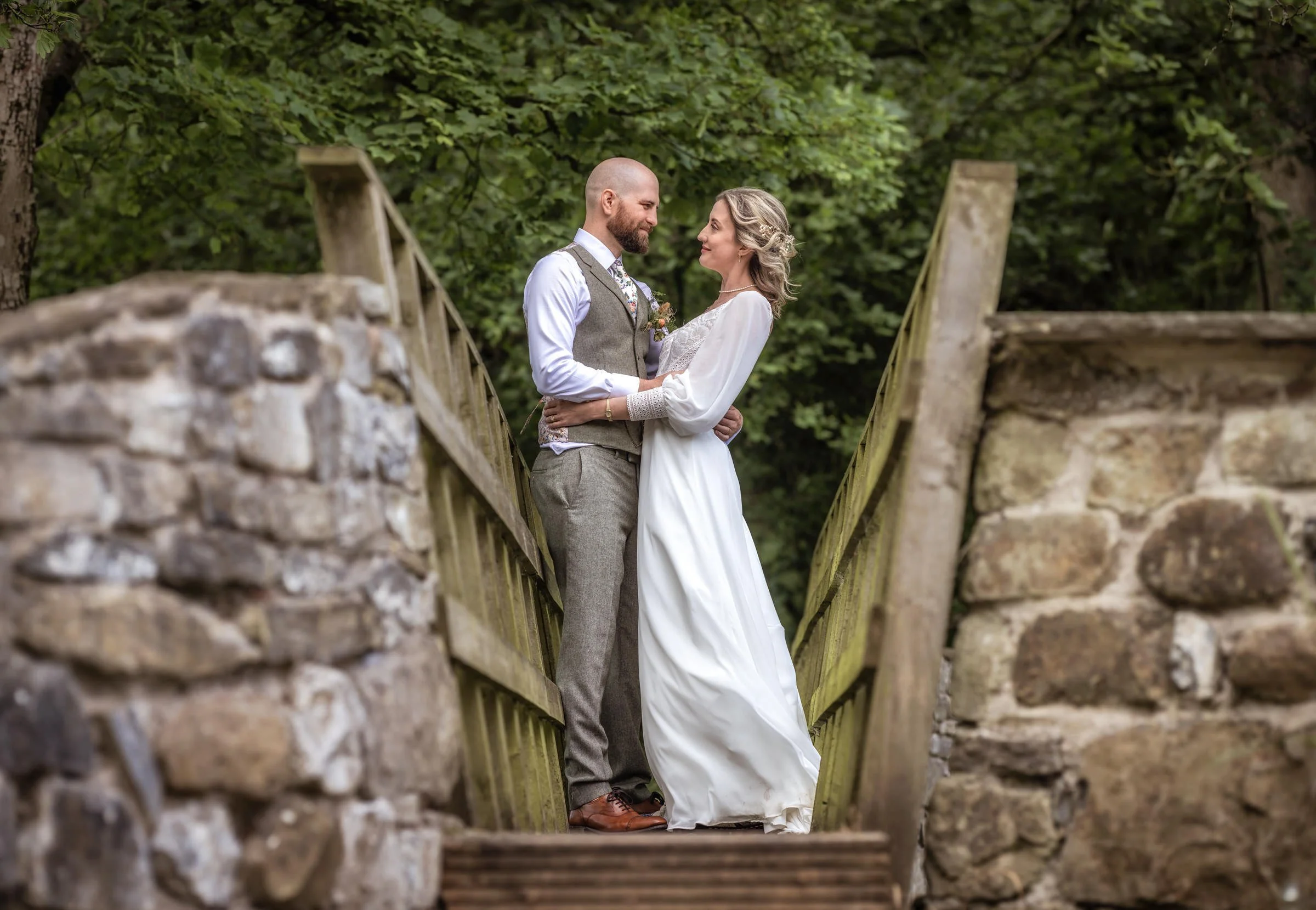 A couple dressed in vintage-style wedding attire standing close on a small wooden bridge surrounded by lush green trees.