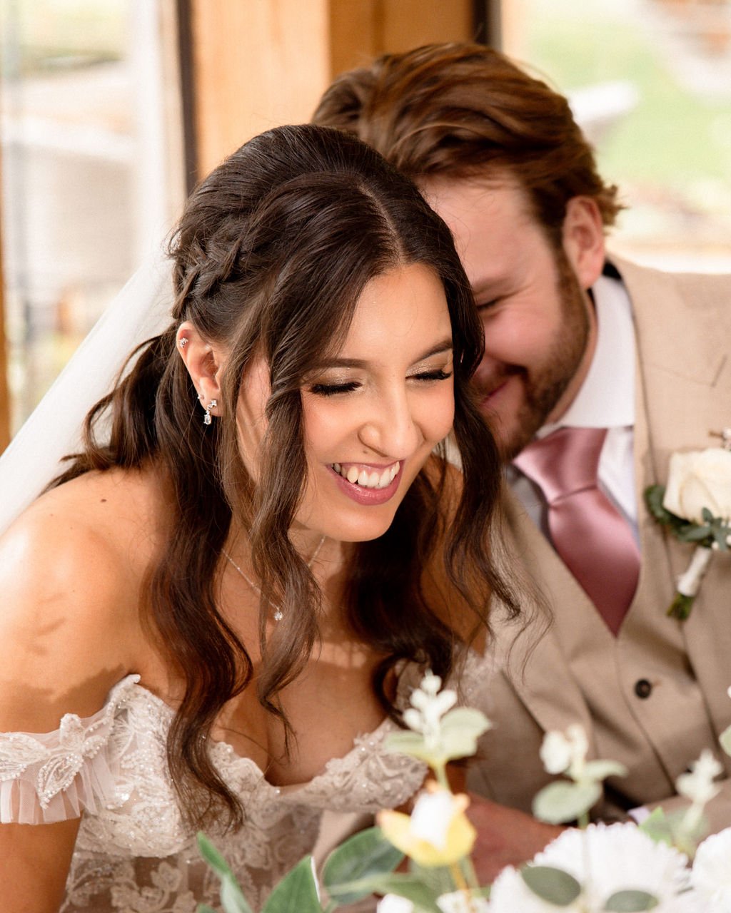 A smiling bride with long brown hair and a groom with short brown hair and a beard, both leaning close together, at a wedding celebration.