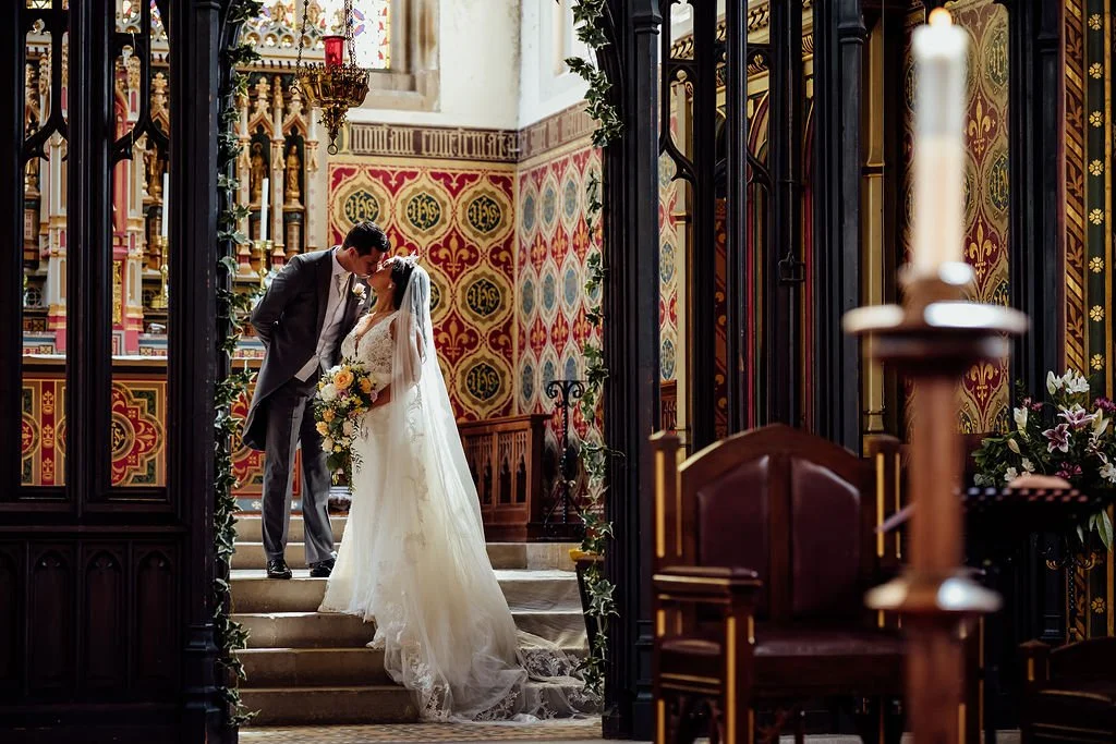 A bride and groom share a kiss on the stairs inside a church, wedding decor with floral arrangements around them, and ornate wall patterns in the background.