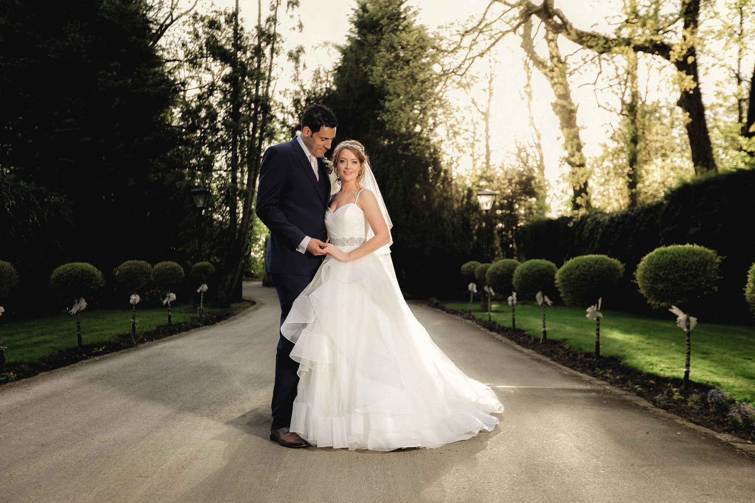 A newlywed couple in wedding attire standing on a tree-lined driveway, with the bride in a white wedding gown and veil, and the groom in a dark suit, holding hands and looking at each other, backlit by the setting sun.