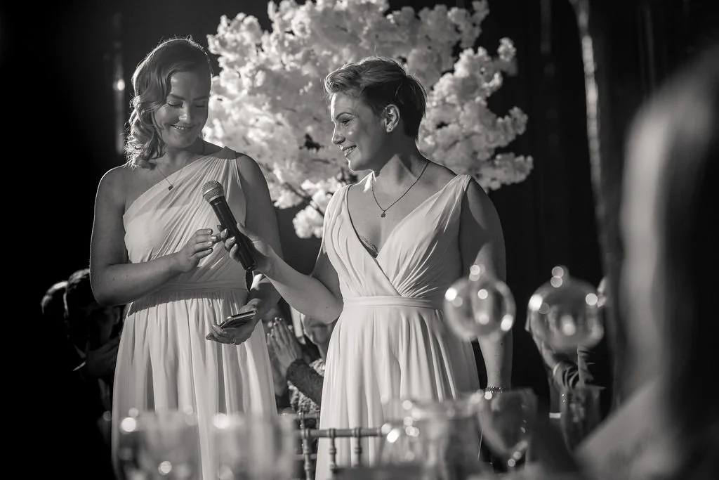 Two women in elegant dresses sharing a moment during a celebration, with floral decorations in the background.