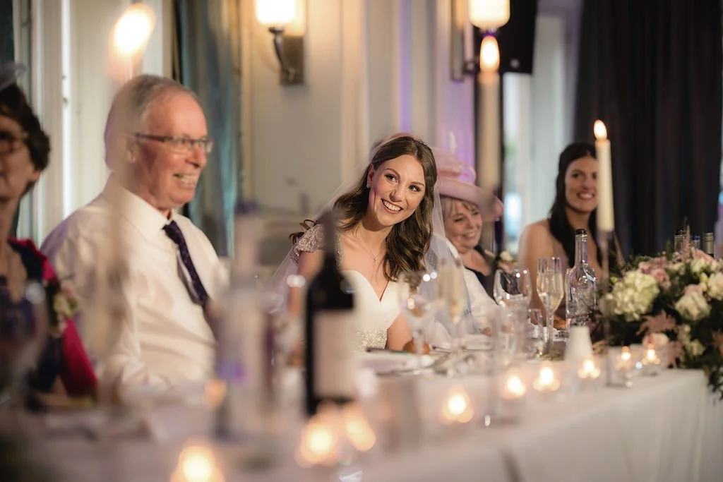 Bride smiling at a wedding reception seated at a decorated table with family and friends, lit candles, and floral arrangements.