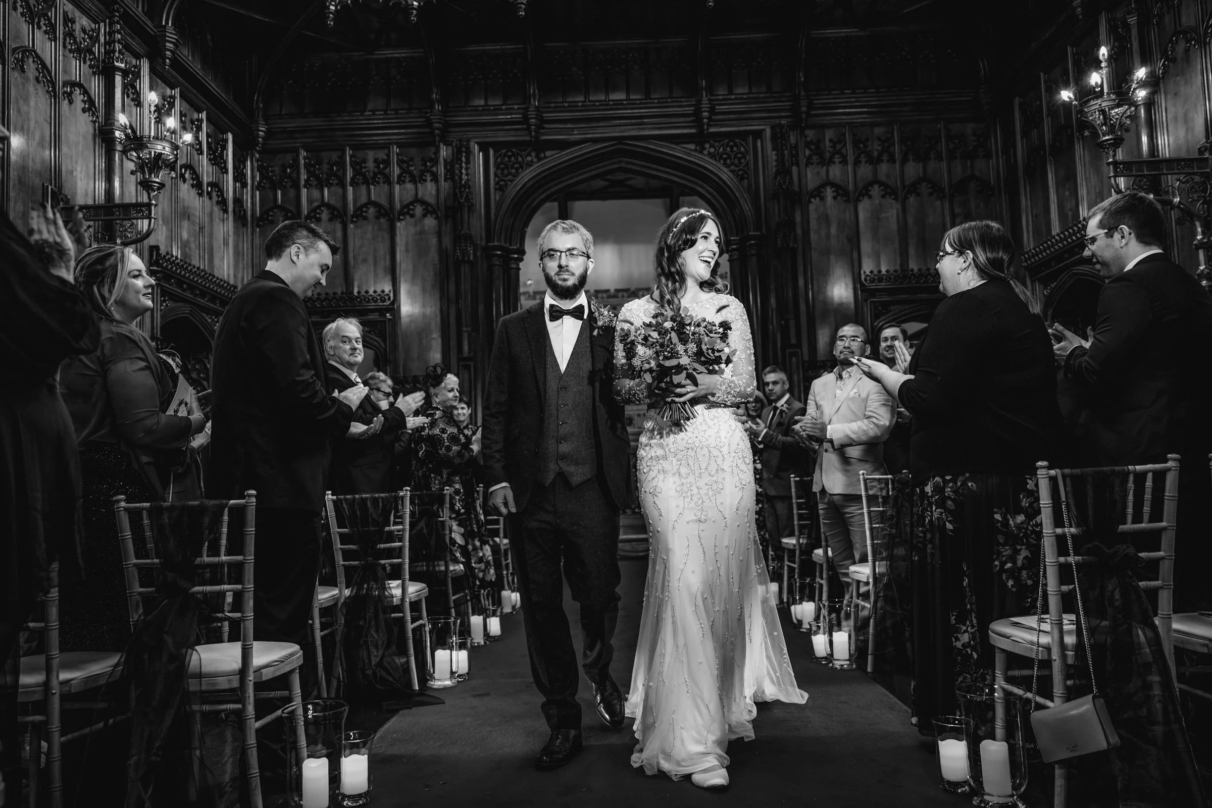 A black and white photo of a wedding ceremony with a bride and groom walking down the aisle, surrounded by seated and standing guests in a grand, wood-paneled room with candles on the floor.