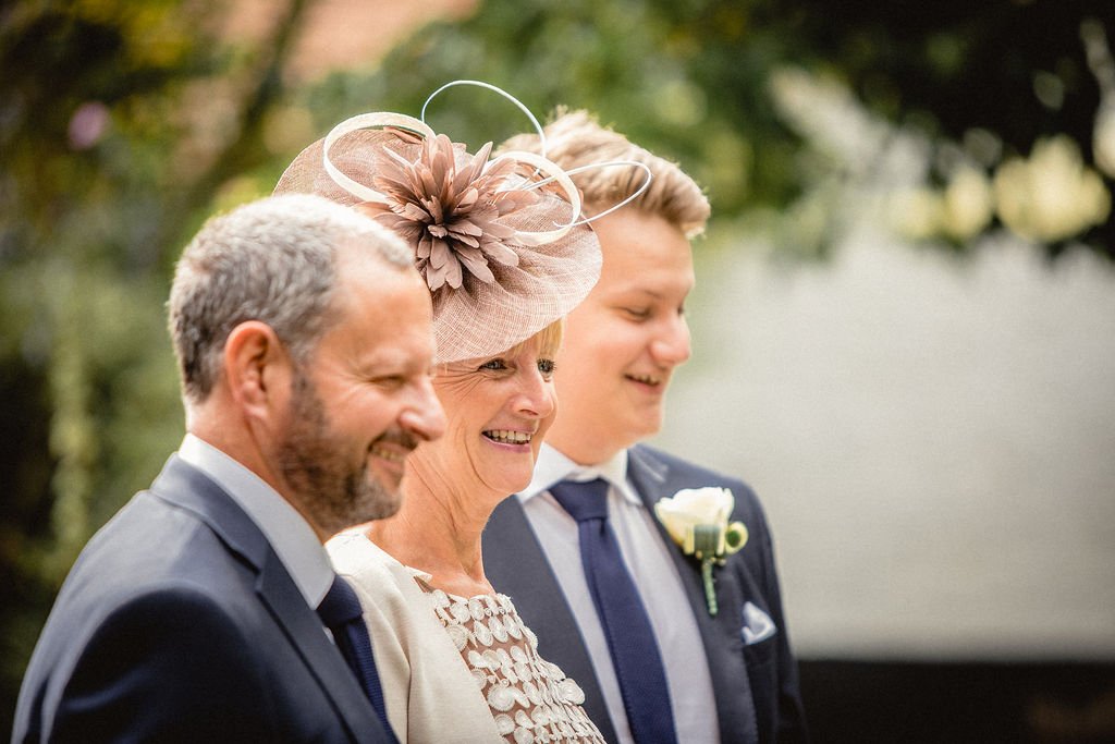 A woman in a pink hat with a large flower and metal accents stands between two men outdoors, all smiling with eyes closed, dressed in formal attire, with blurred greenery in the background.