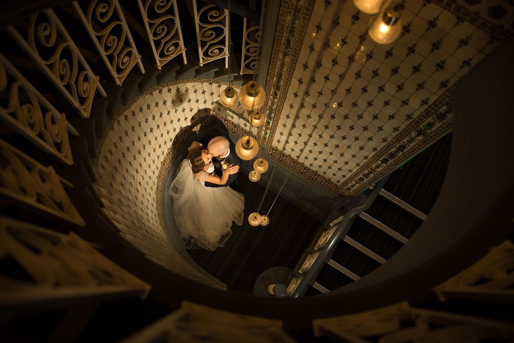 A bride and groom seated together on a staircase, seen from above, under hanging light fixtures, with ornate decor and patterned walls.