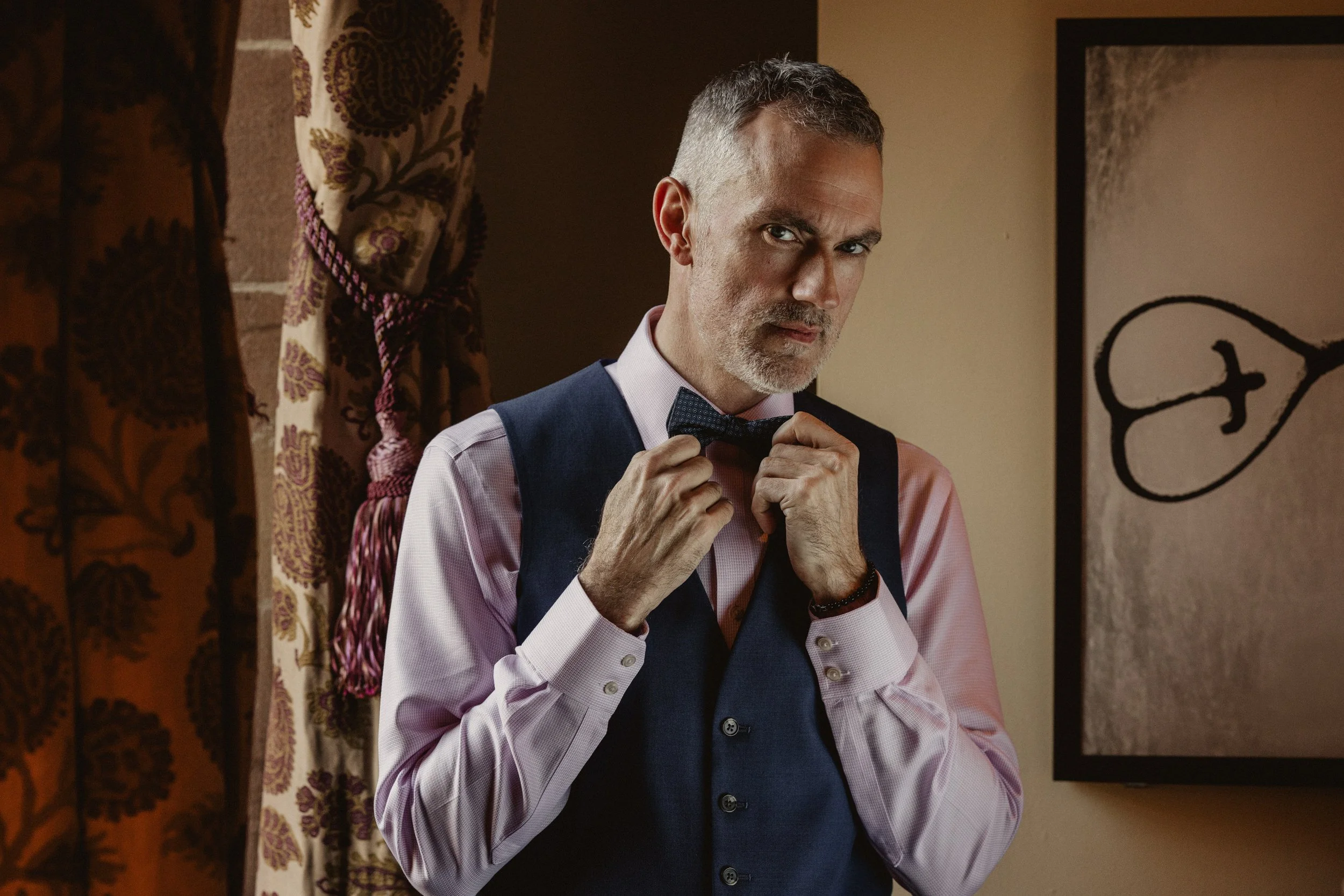 A middle-aged man with gray hair and a beard adjusting a bow tie, dressed in a pink shirt and a dark vest, standing indoors.