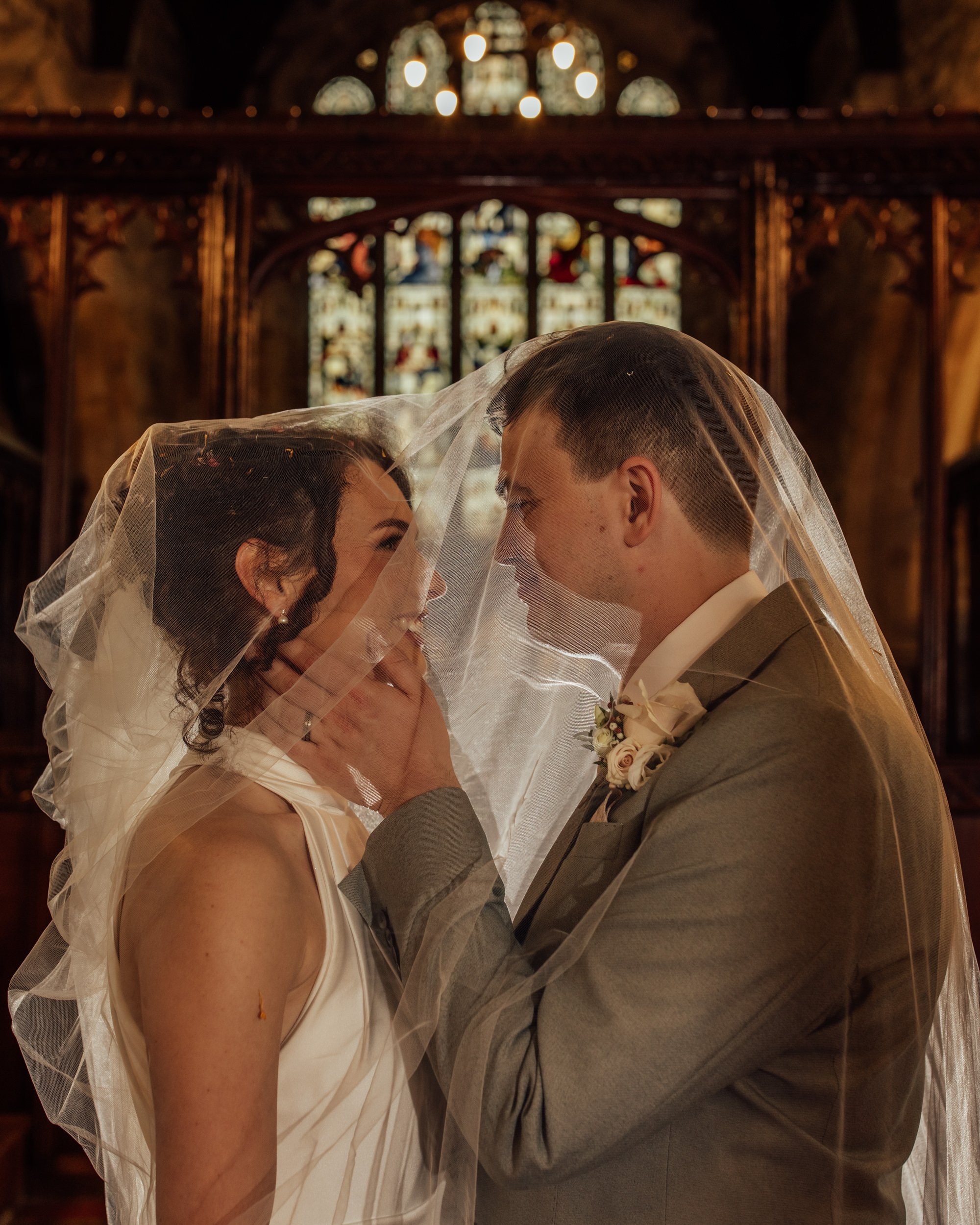 A bride and groom face each other inside a church, touching foreheads beneath a wedding veil, with stained glass windows and ornate woodwork in the background.
