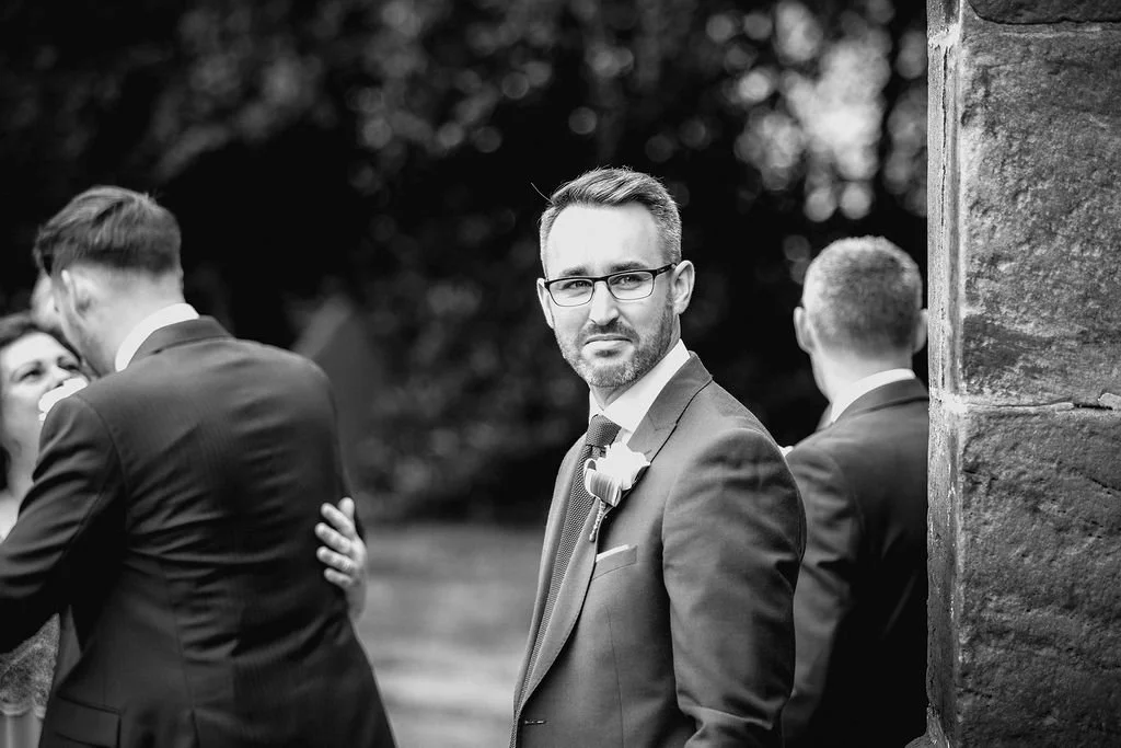 A man in a suit and glasses standing outdoors next to a stone wall, with other people in suits in the background at a formal event, likely a wedding.
