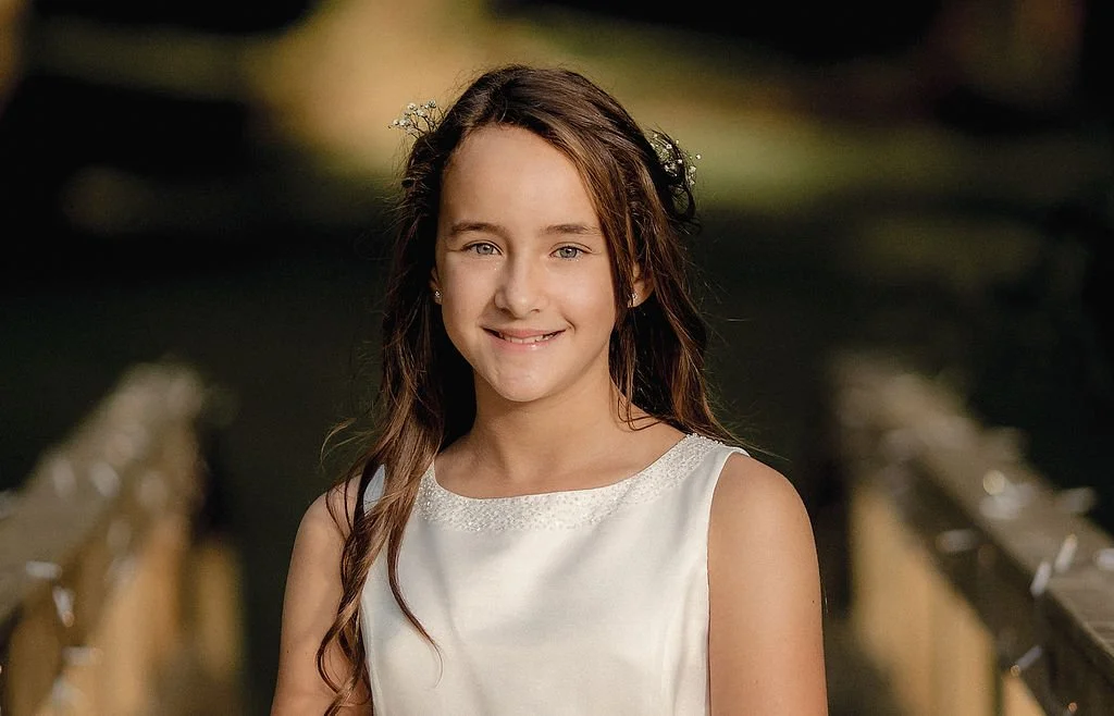 A young girl with long brown hair, wearing a white sleeveless dress, smiling and standing outdoors on a bridge with blurred greenery in the background.