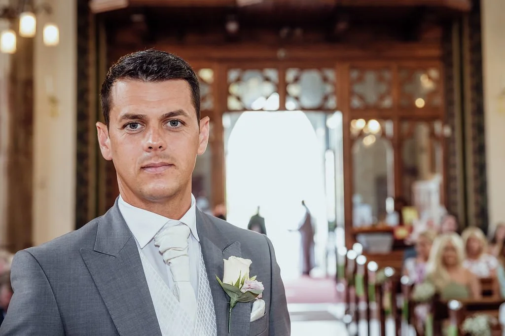 A groom in a gray suit with a white boutonniere at a wedding ceremony inside a church.