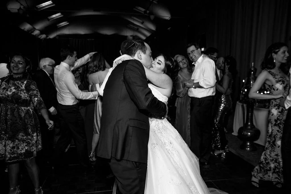 A black and white photo of a wedding reception dance floor with a newlywed couple embracing and dancing, surrounded by guests enjoying the celebration.
