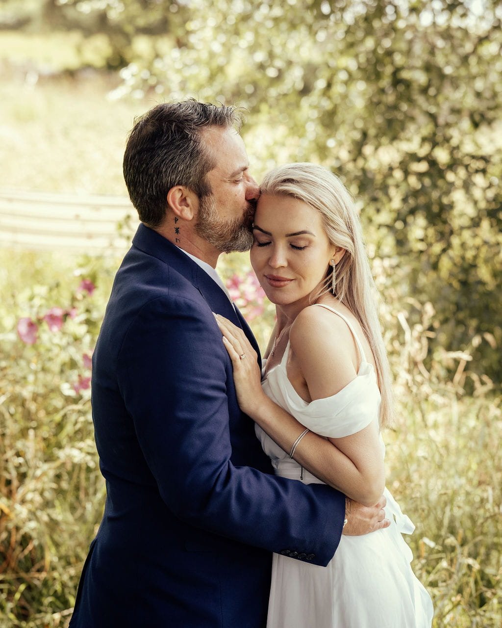 A man with gray hair and beard kissing a woman with long blonde hair on her forehead in a garden setting during daytime.