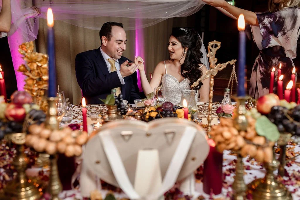Bride and groom sitting at decorated wedding reception table, smiling and holding hands, surrounded by candles, flowers, grapes, and guests.