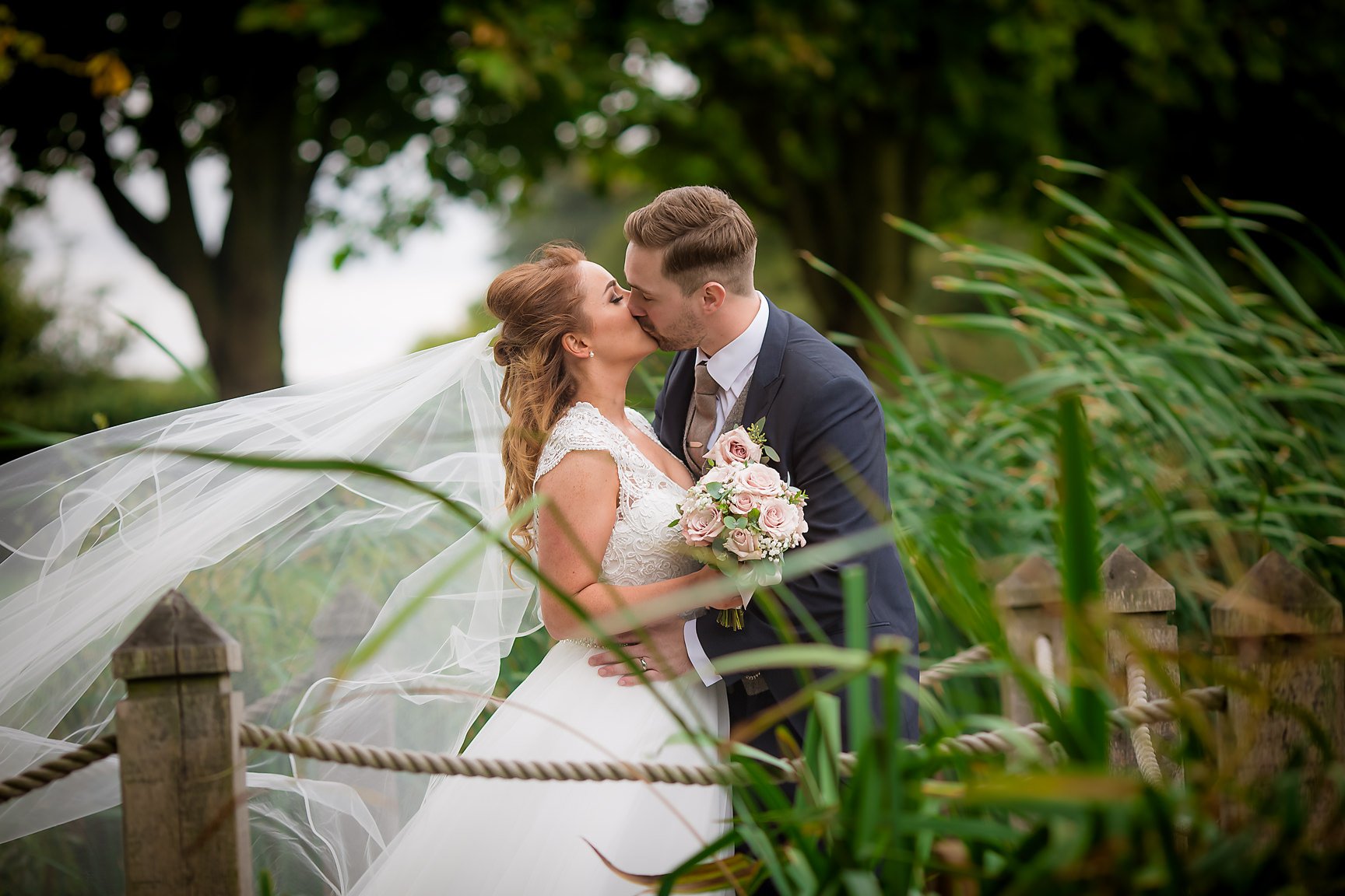 Bride holding a white, green and pink bouquet, kissing her groom in outdoor setting with wind blowing her veil and grass