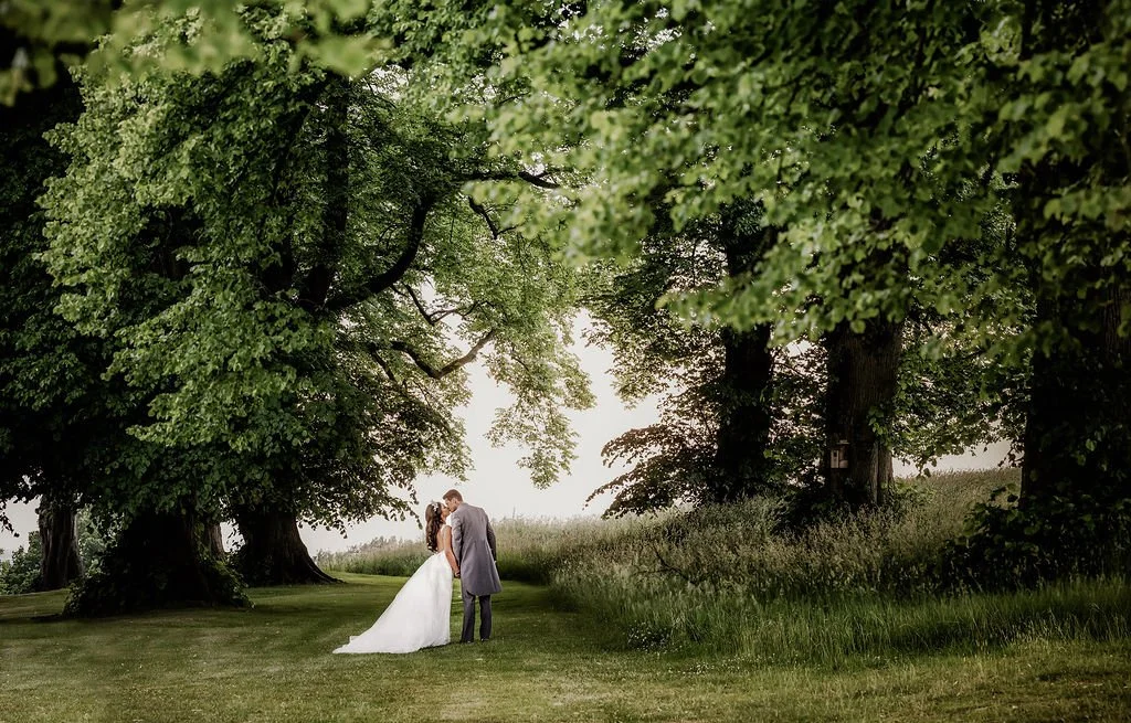 A wedding couple sharing a kiss under large green trees in a grassy field.
