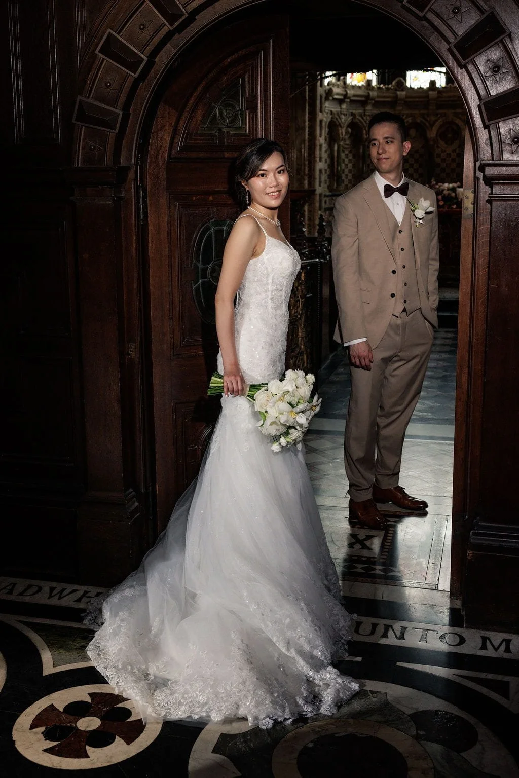 A bride and groom standing in a church, the bride holding a bouquet of white flowers, wearing a white wedding gown, and the groom in a tan suit with a bowtie, behind a wooden archway.