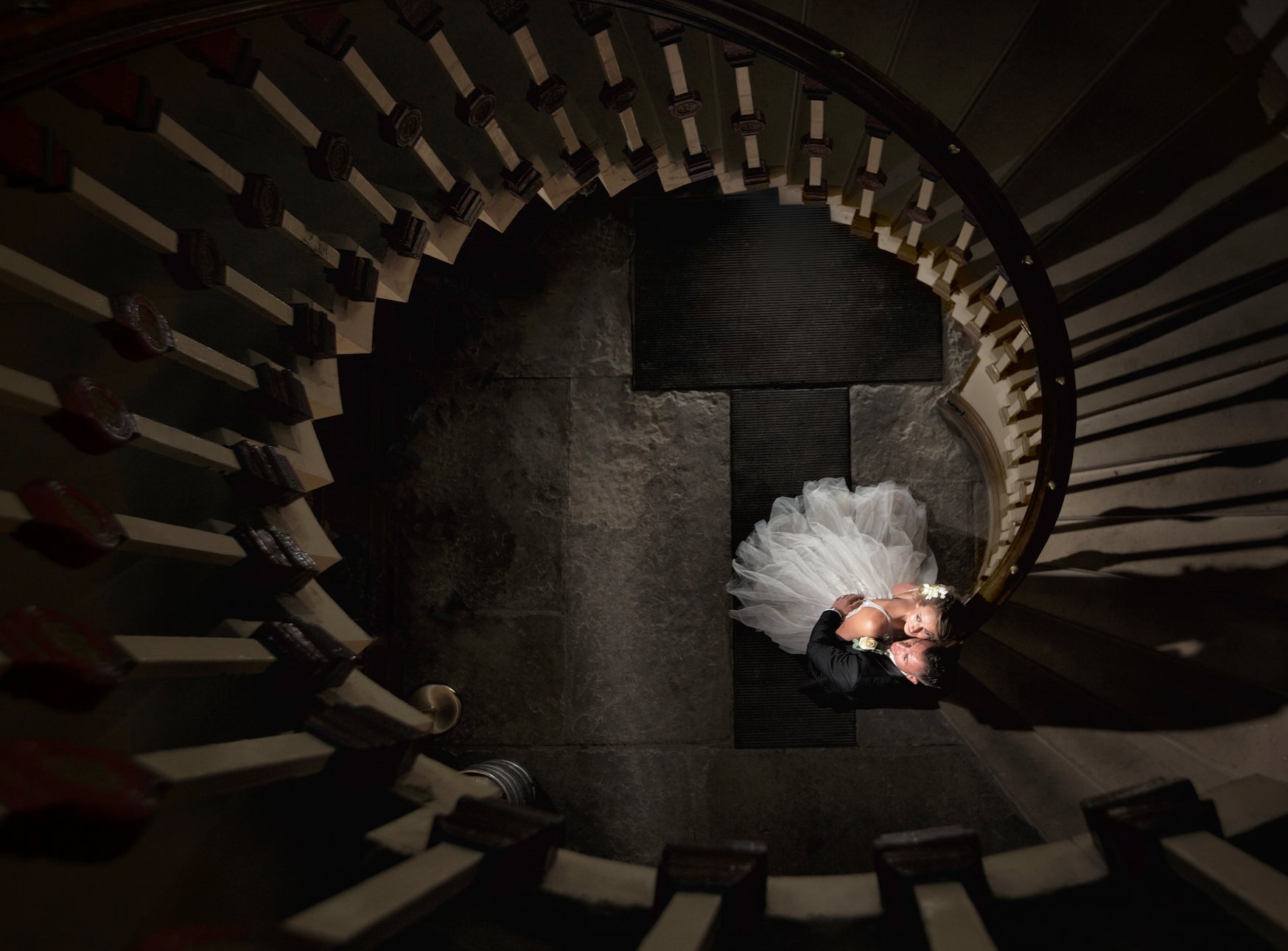 Wedding couple kissing on a staircase with a curved railing, seen from above. The bride is in a white wedding dress and the groom in a black suit. The photo is taken indoors on a dark stone floor.