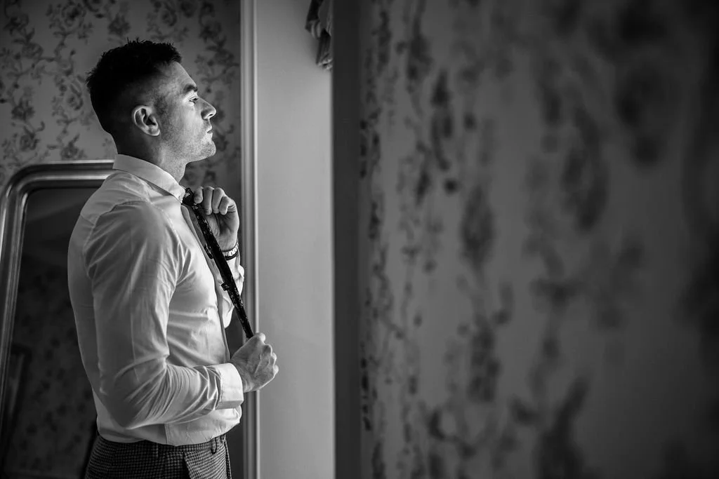 A young man in a white dress shirt with a tie, standing in front of a mirror, holding the tie with one hand, looking thoughtfully.