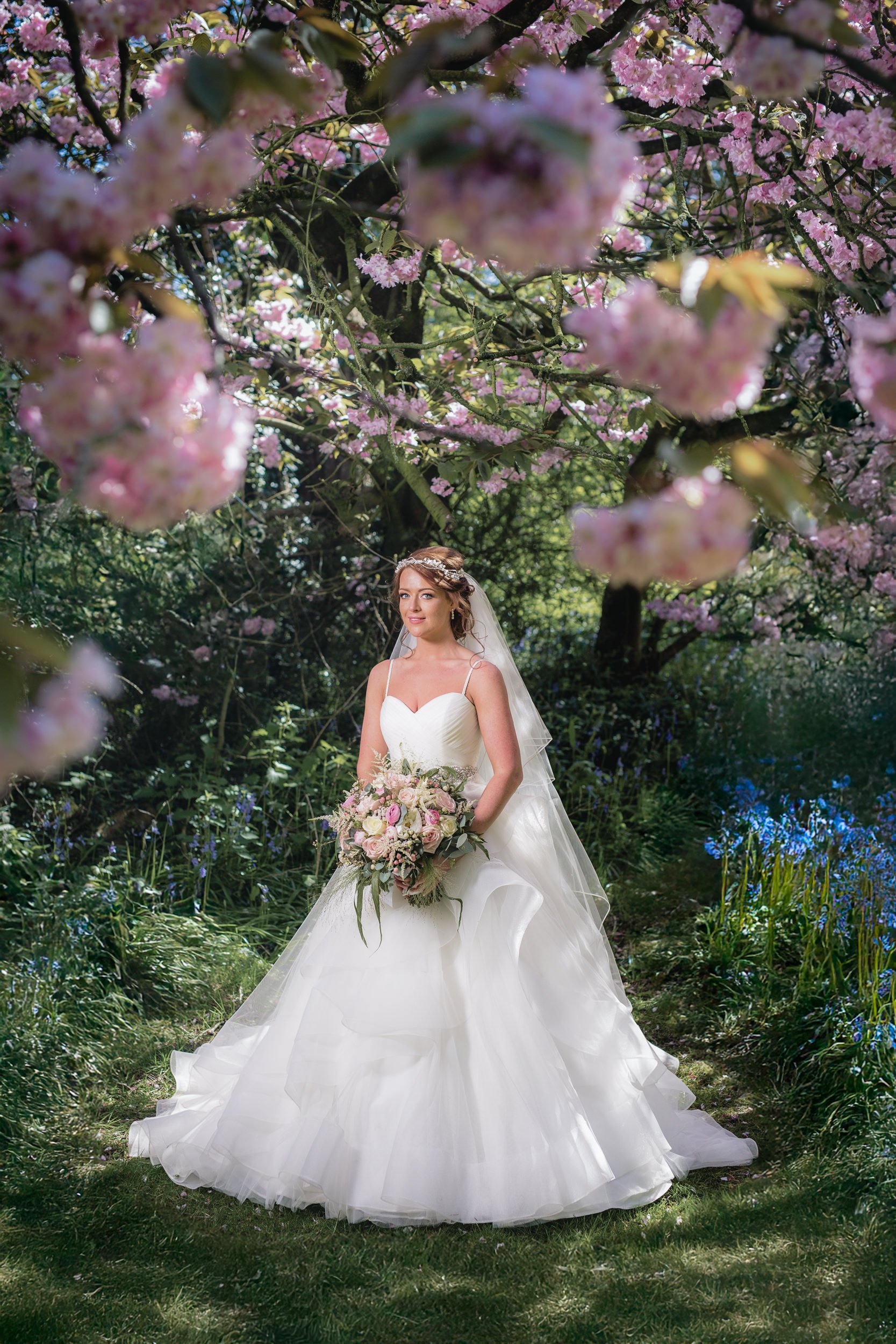 A bride in a white wedding gown holding a bouquet stands in a lush garden surrounded by blooming pink and purple flowers.