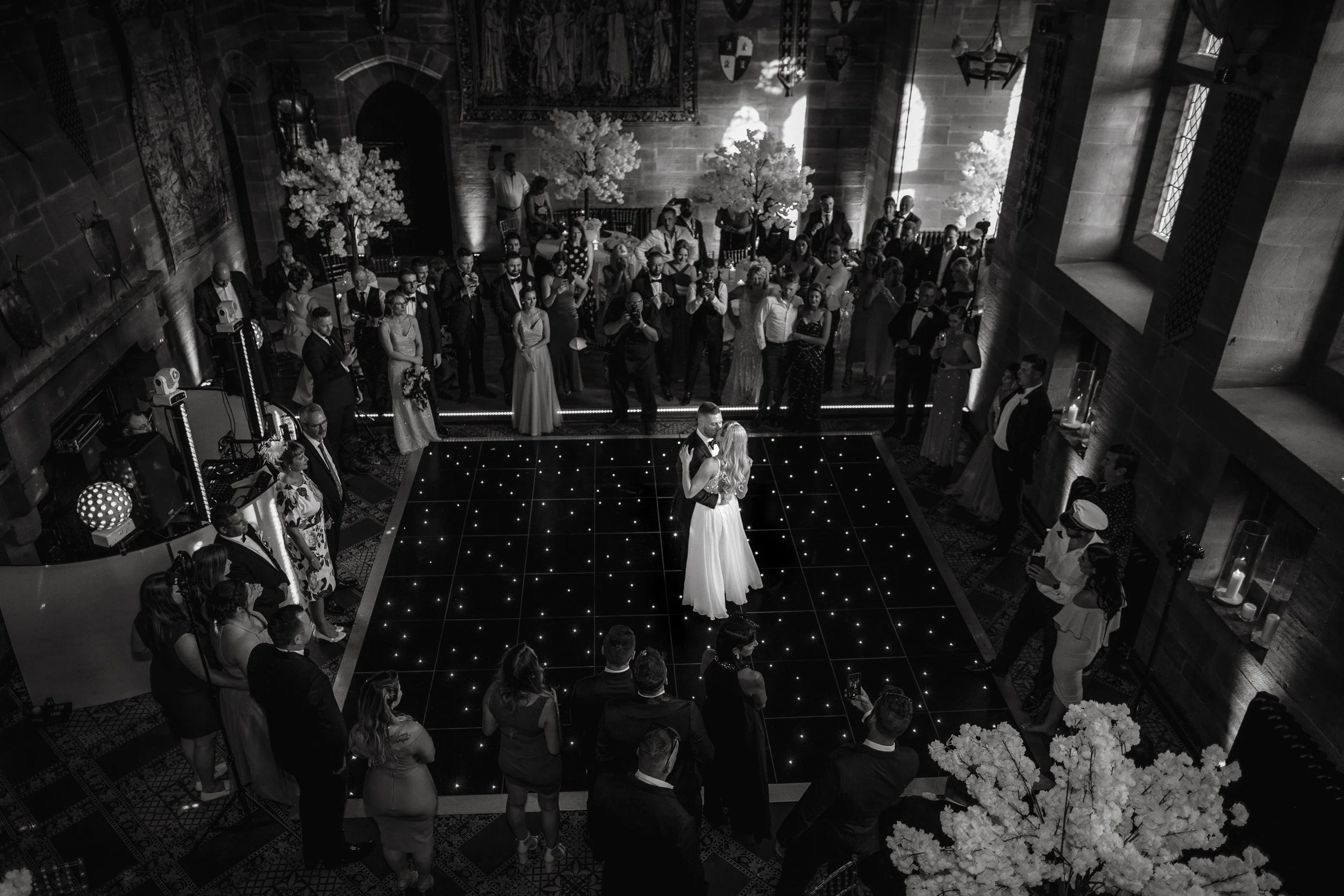 A couple is dancing together on a lit-up dance floor at their wedding reception, surrounded by wedding guests in an elegant, candle-lit banquet hall with large windows, floral arrangements, and decorative trees.