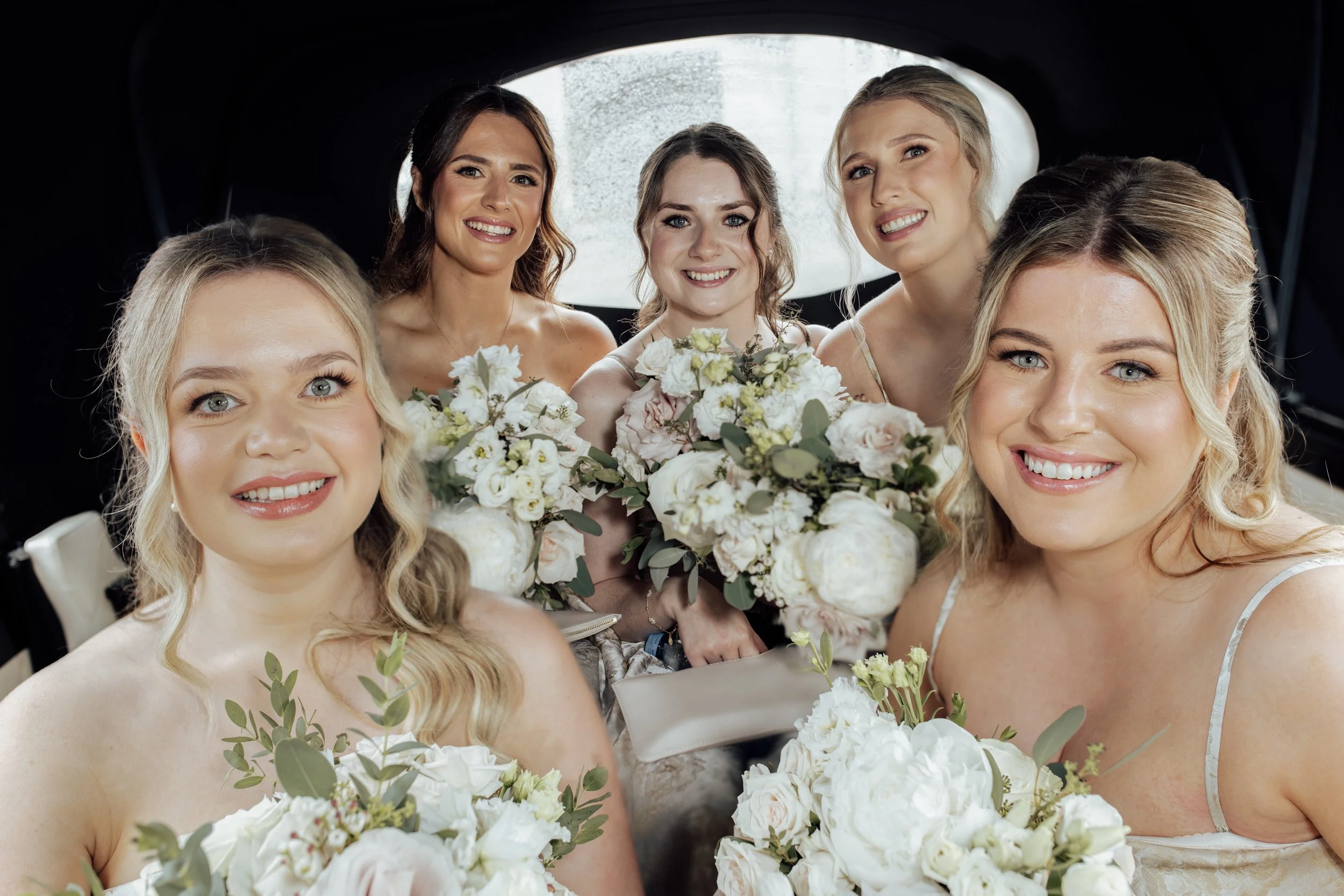 A group of women, including a bride, smiling and holding bouquets of white flowers during a wedding celebration inside a vehicle.