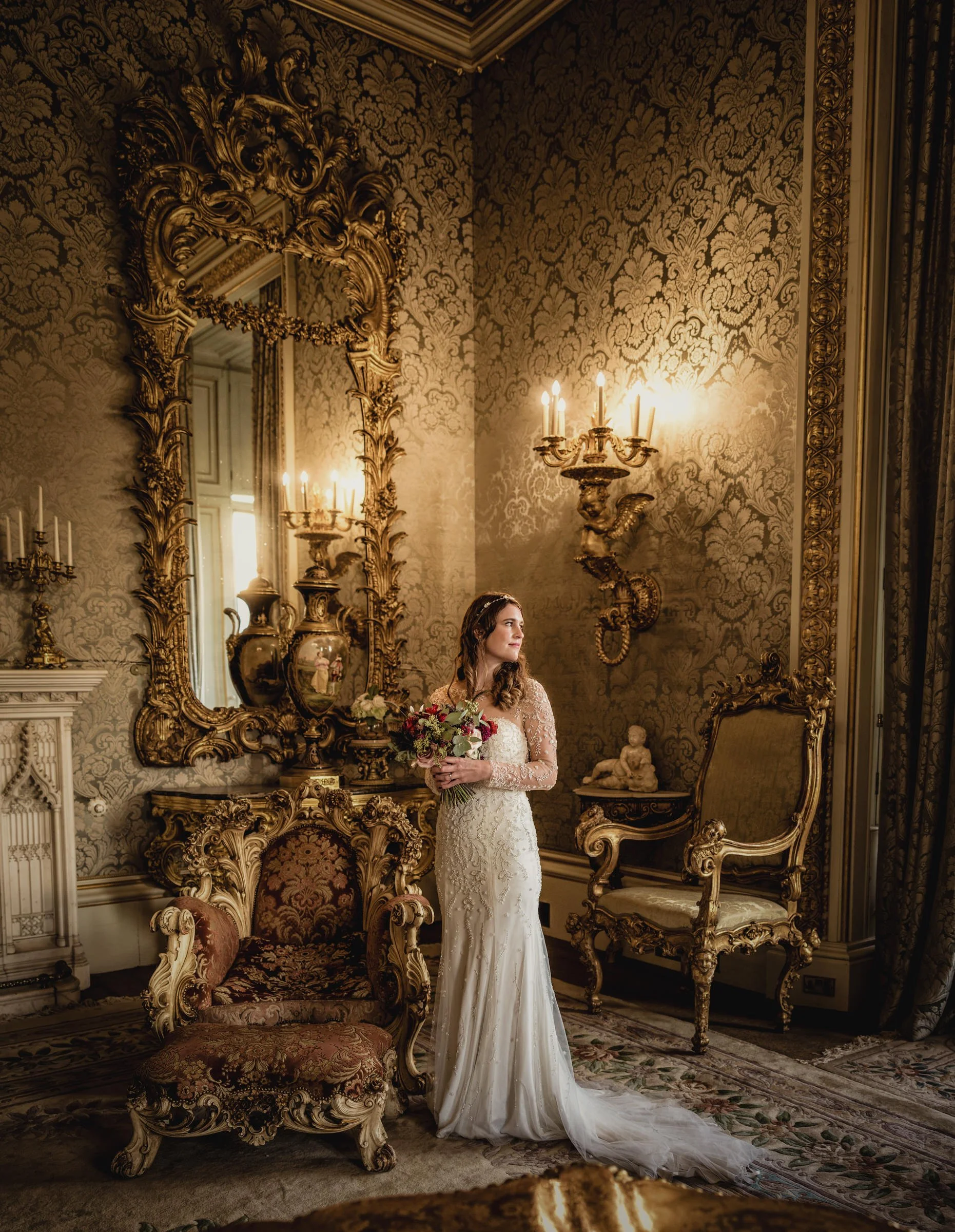 A bride in a lace wedding dress holding a bouquet standing in an ornate, vintage-style room with gilded furniture, a large mirror, and elaborate wallpaper.