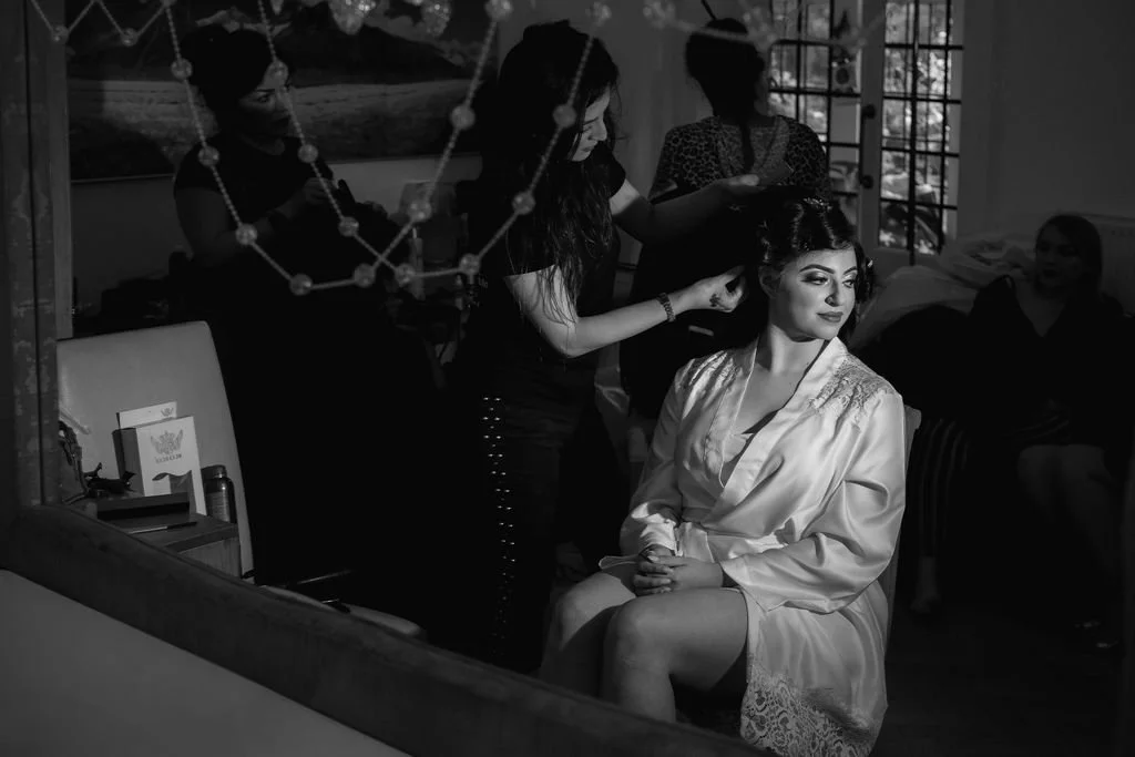 A woman getting her hair styled by a hairstylist while sitting in a chair, with other people in the background in a room with a window and decorative string lights.