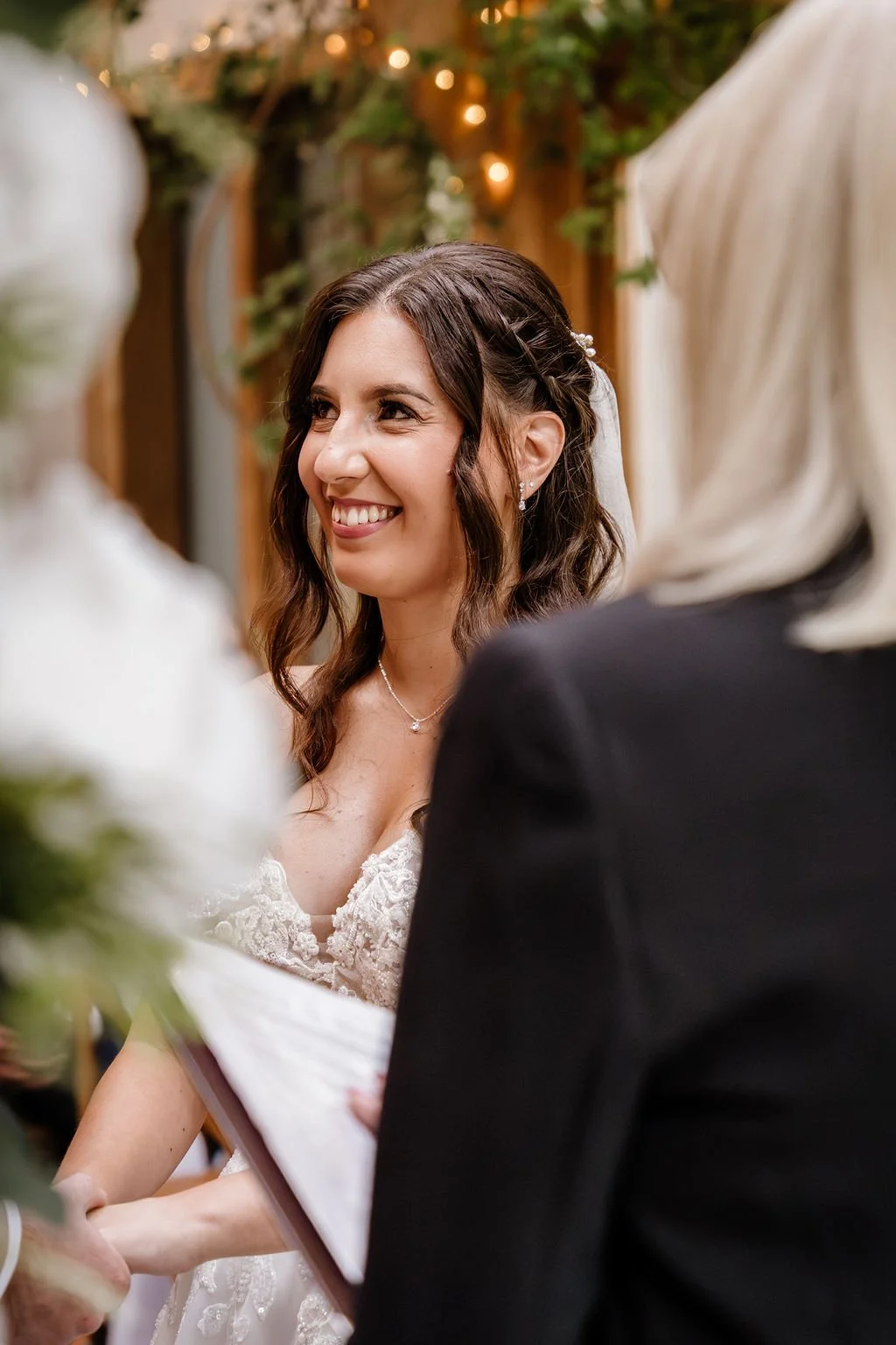 A bride in a wedding dress smiling during her wedding ceremony, with a person holding a book or notes. The background features greenery and warm lighting, and the bride has dark wavy hair, earrings, and a necklace.