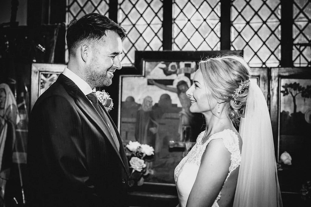 A bride and groom exchanging vows or exchanging looks at their wedding ceremony, with a church interior and stained glass windows in the background.