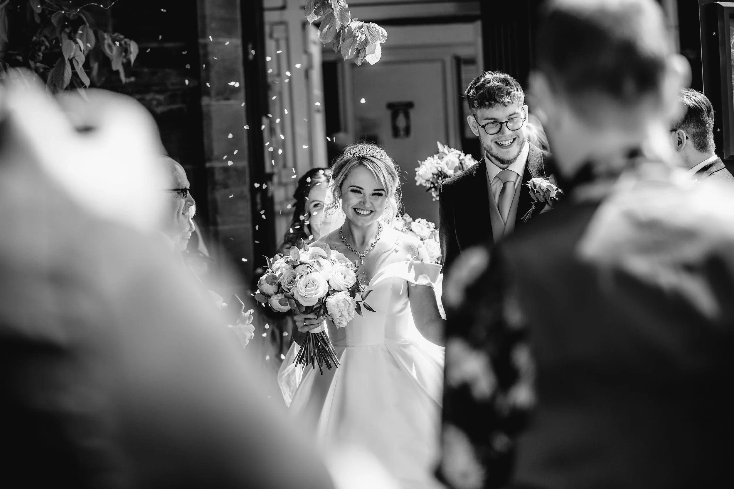 Black and white photo of a wedding celebration with a smiling bride holding a bouquet of flowers, surrounded by cheerful guests.